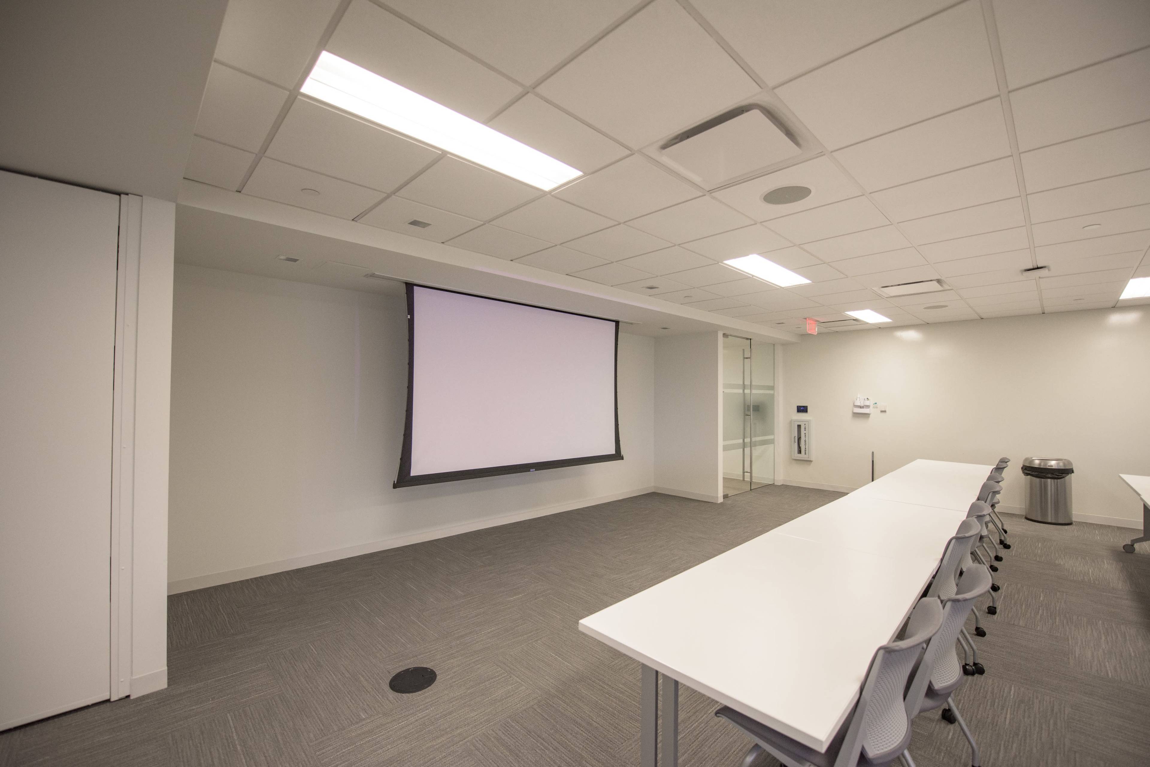 A vacant conference room with a projector screen on one wall and a long white table with chairs arranged alongside it.