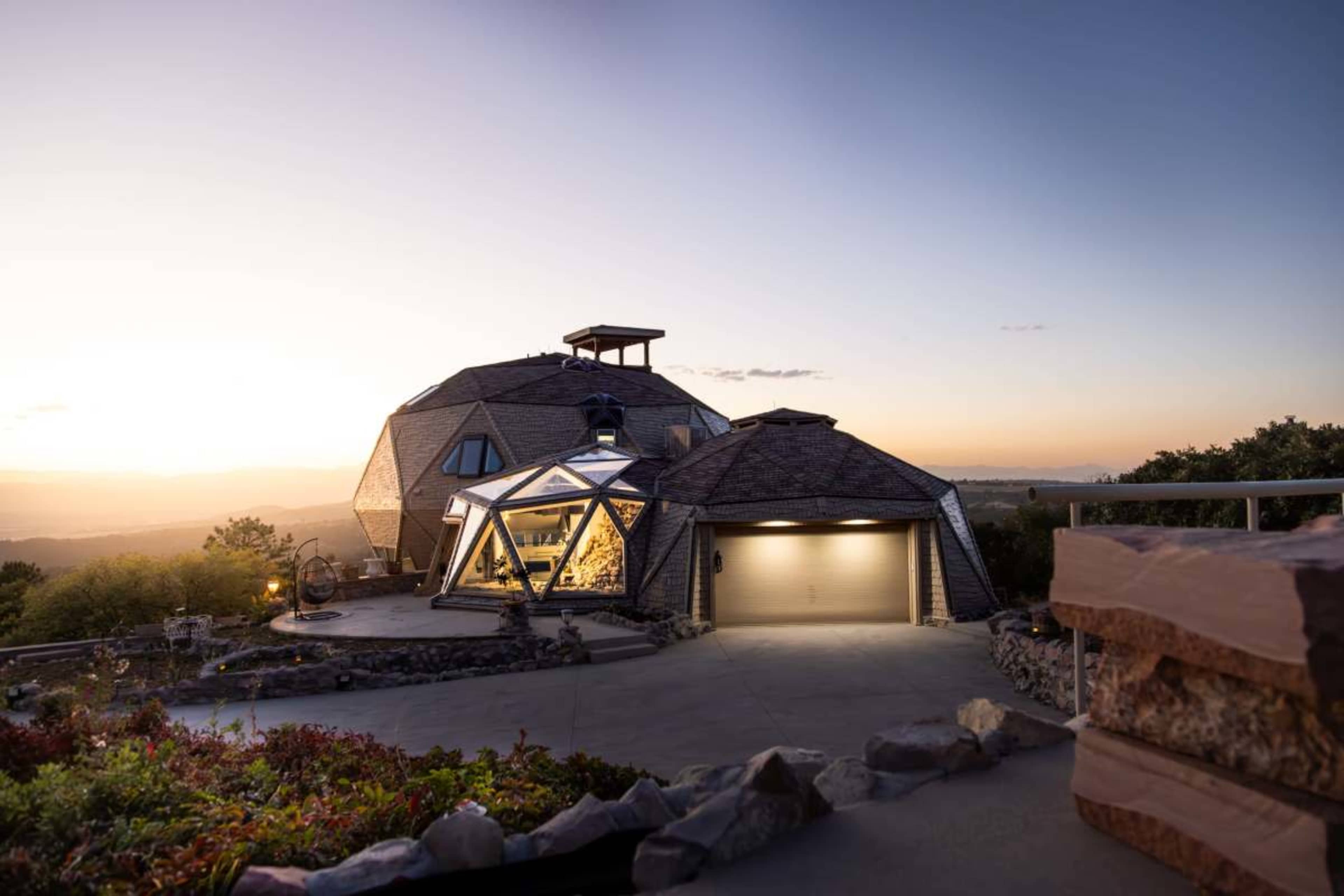 A geodesic dome house with large glass windows is situated on a hillside at sunset.