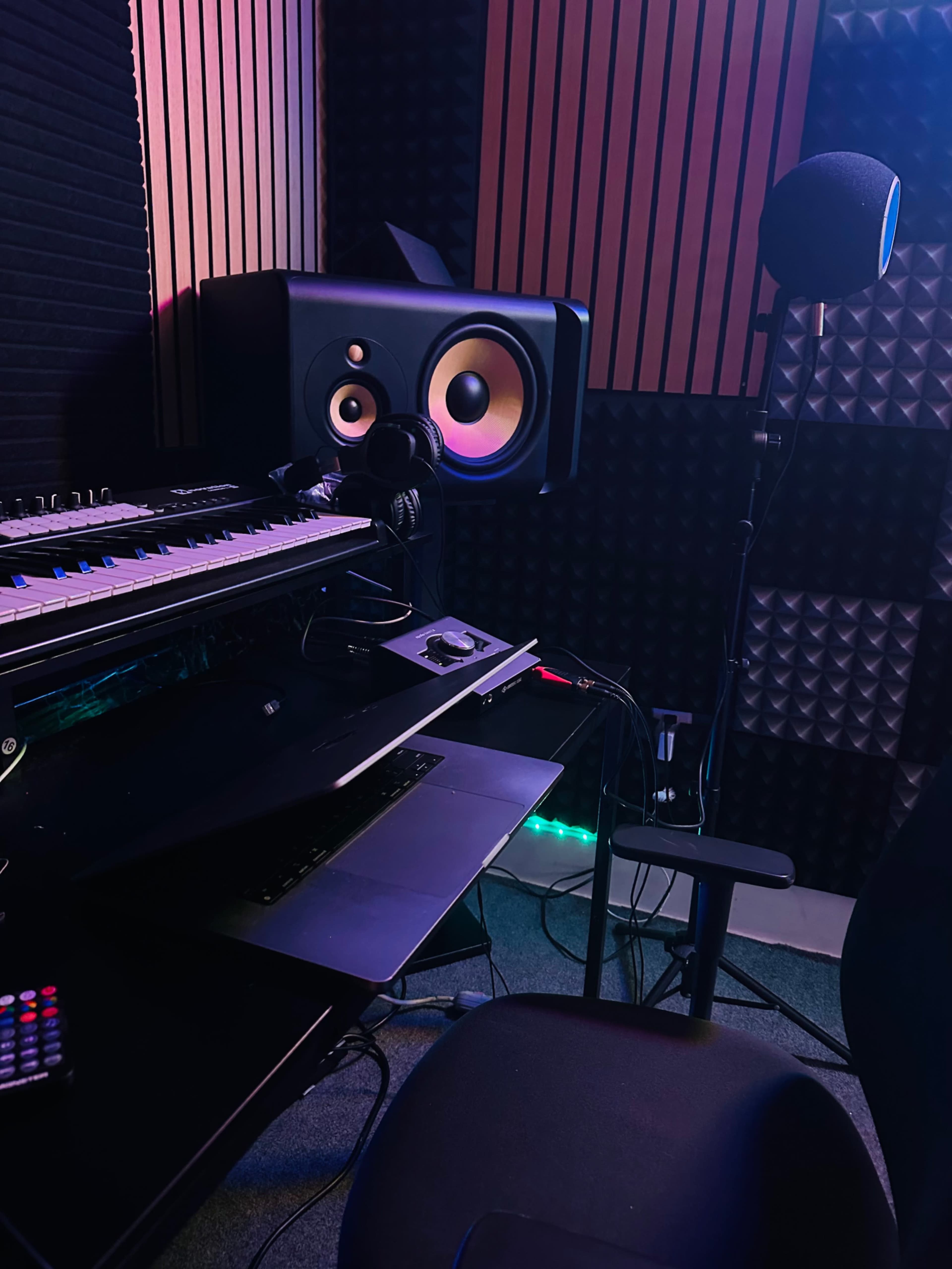 The image shows a music recording studio setup with a keyboard, audio interface, studio monitors, and a microphone stand in a sound-insulated environment.