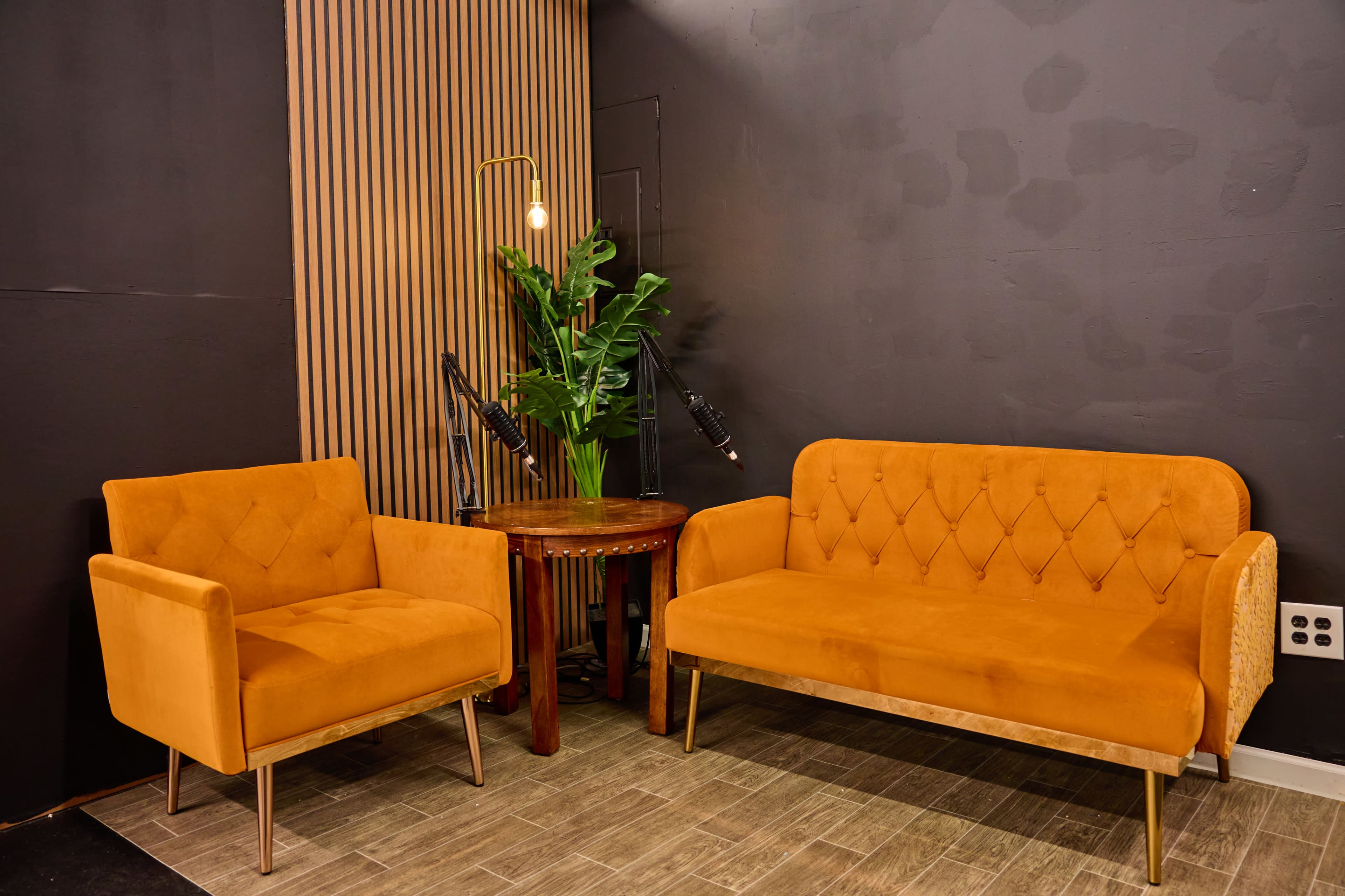 A cozy seating area with an orange sofa and armchair positioned beside a round wooden side table, accompanied by a potted plant against a dark wall.