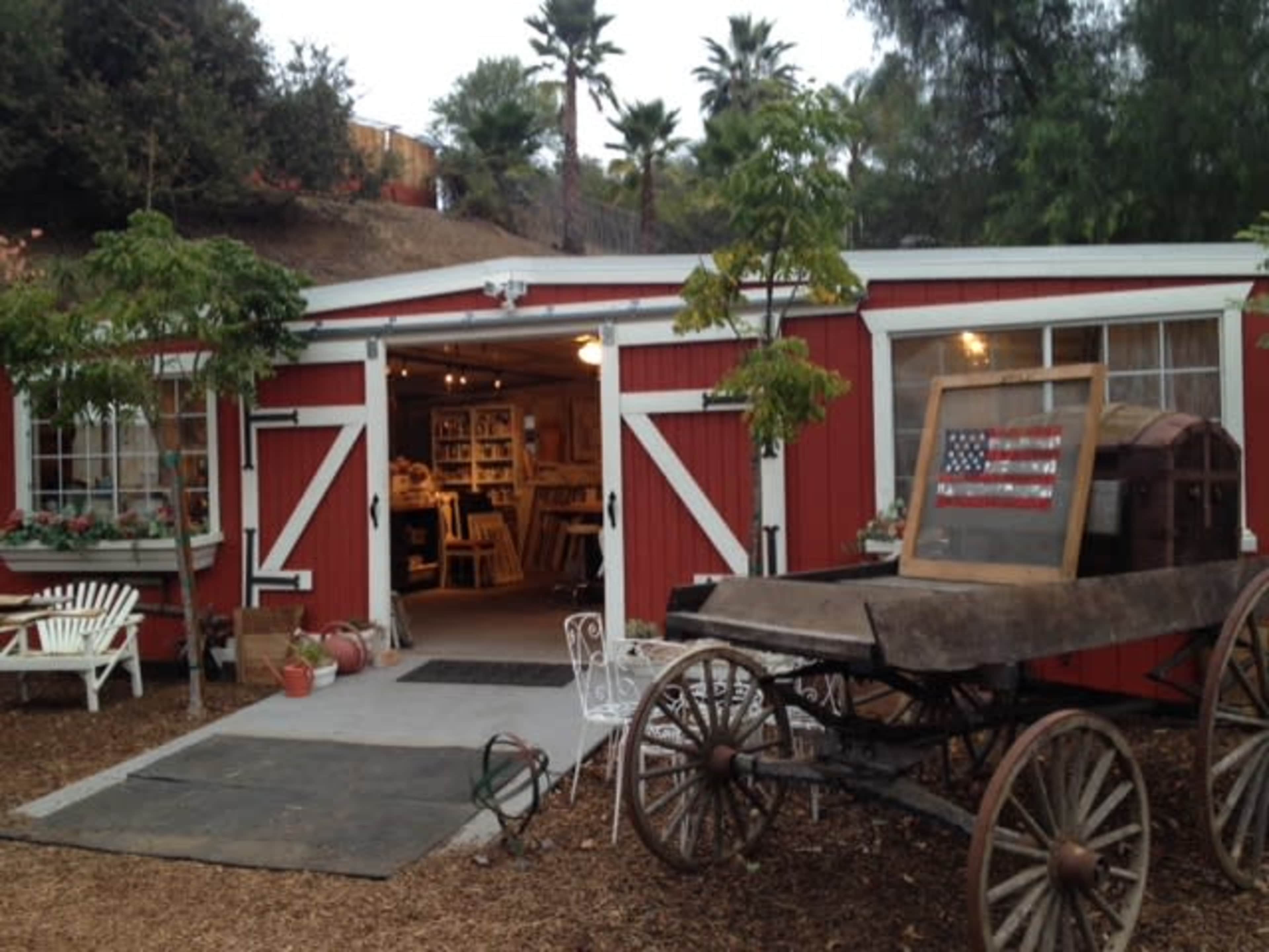 A red barn with white trim and double doors stands next to a vintage wagon, surrounded by potted plants and trees.