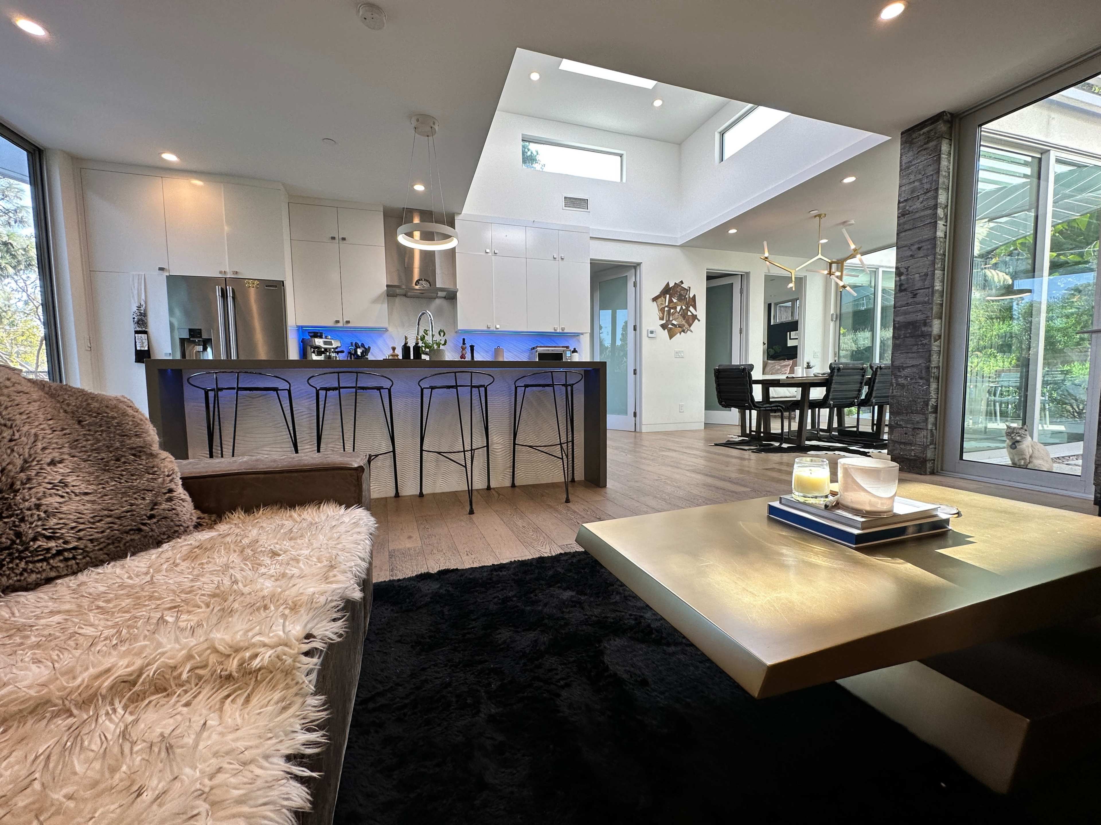 The image shows a modern open-concept living space with a kitchen featuring a large island and bar stools, a dining area, and large windows allowing natural light.