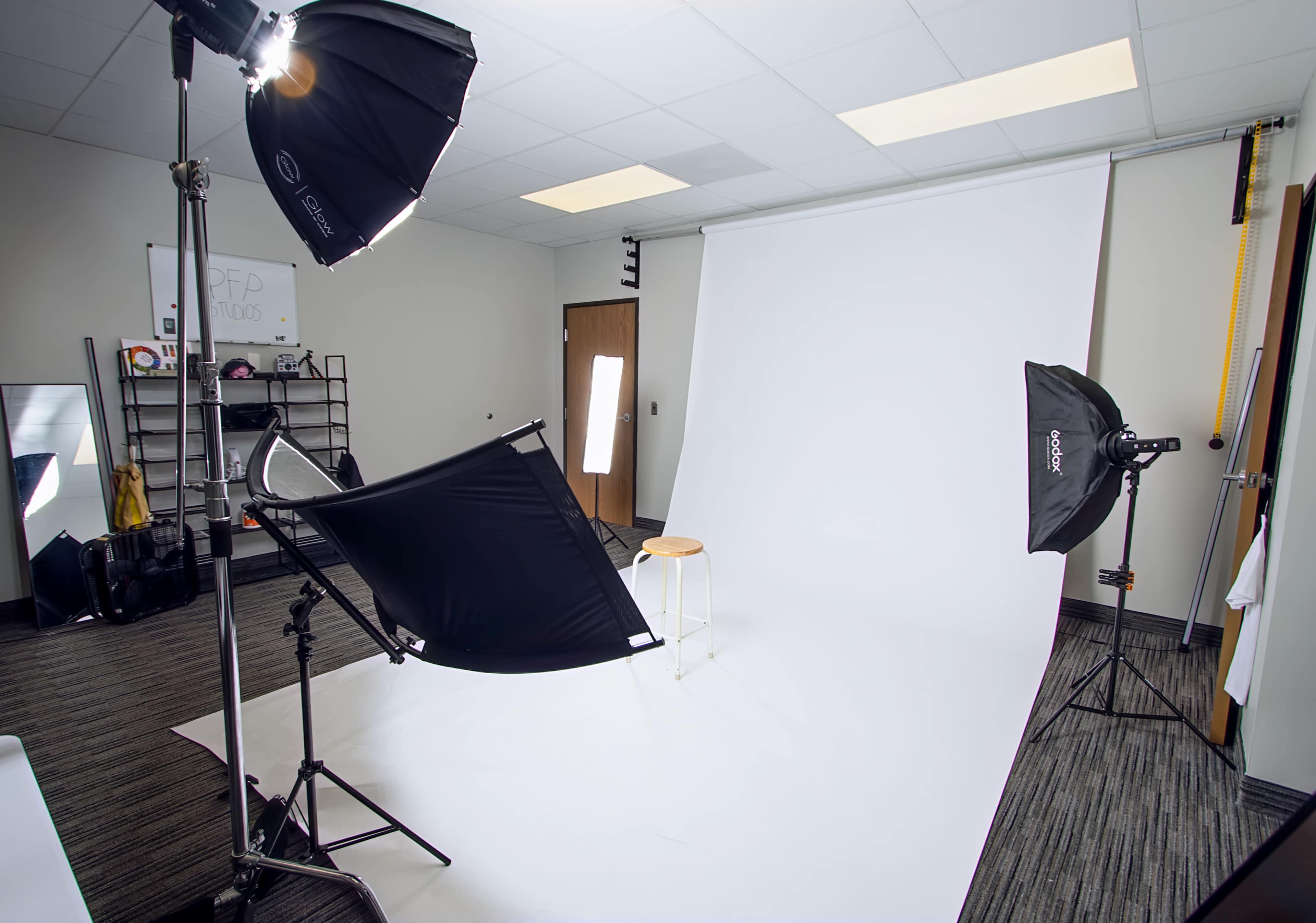 The image shows a photography studio equipped with softbox lights, a white backdrop, and a stool positioned in front of it.