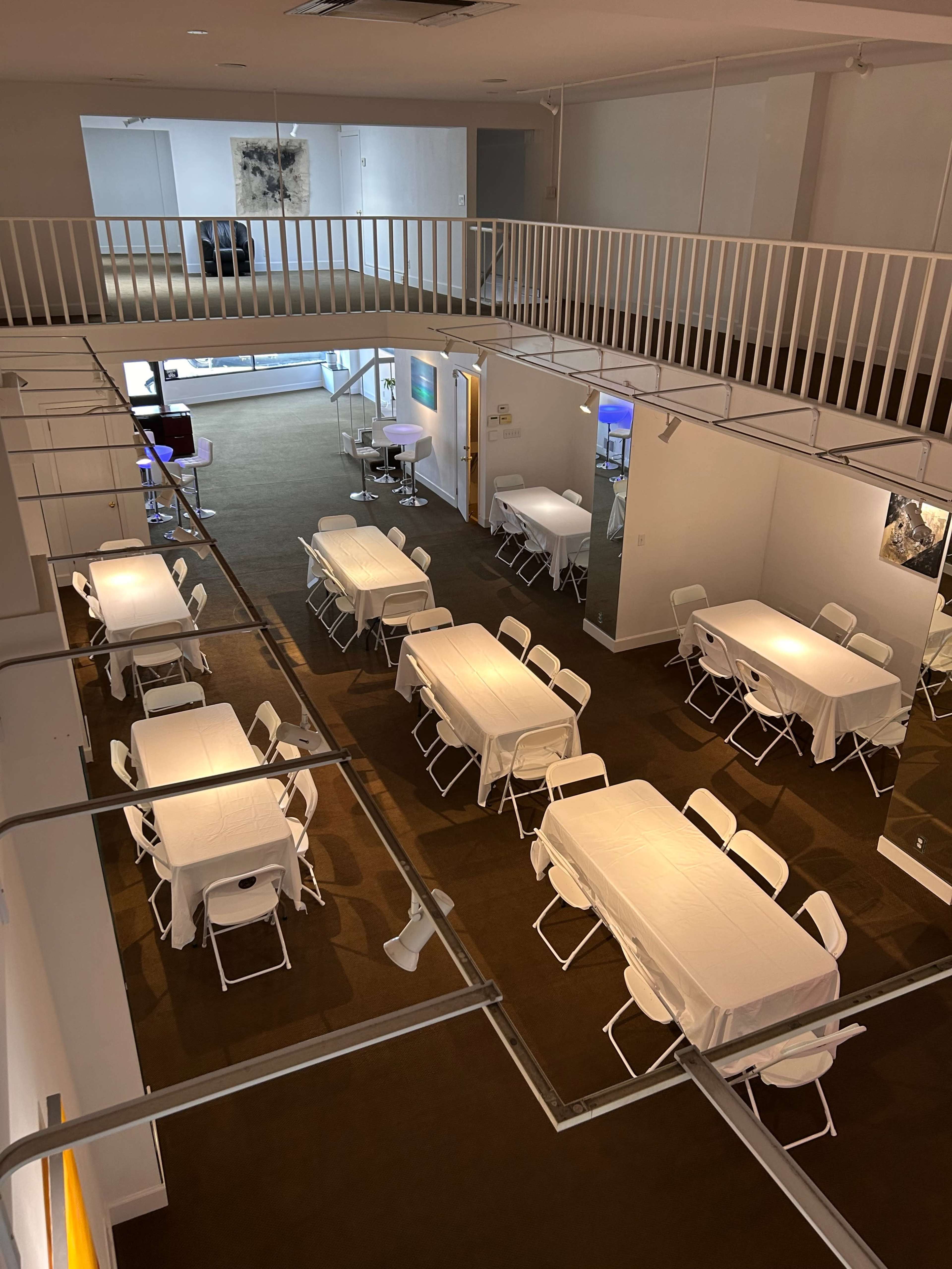 The image shows a spacious, well-lit room with several tables covered in white cloth, arranged in a grid pattern, and a balcony overhead.