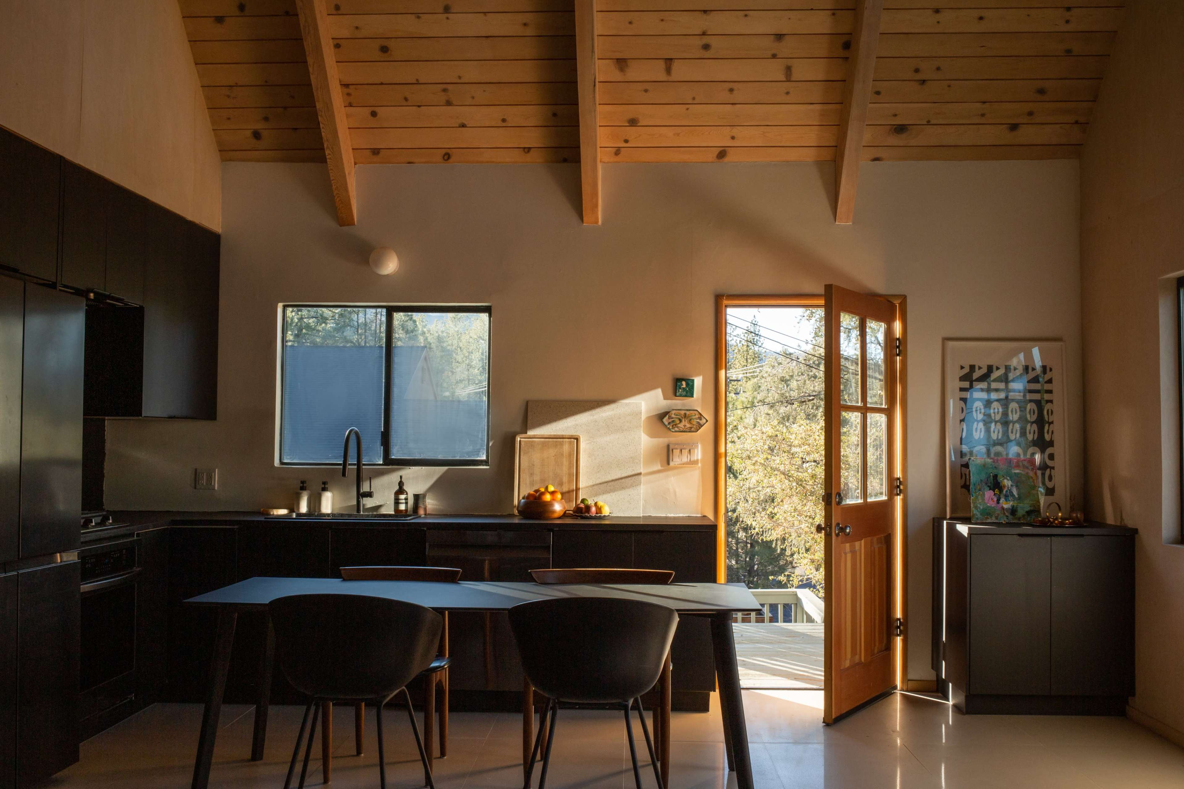 A modern kitchen with wooden beams, a table, and an open door leading to an outdoor area filled with natural light.