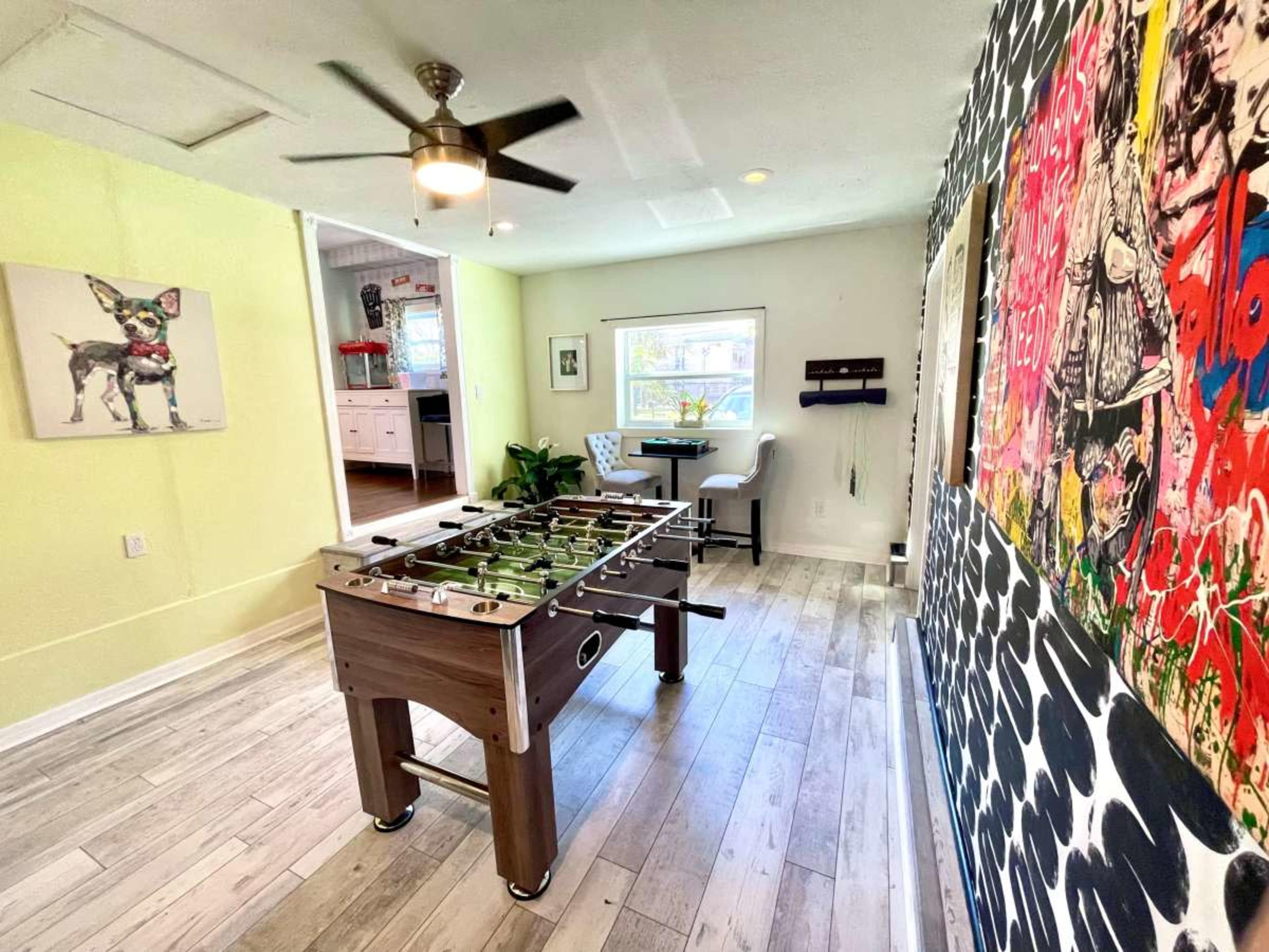 The image shows a recreational room with a foosball table in the center, flanked by a colorful artwork wall and a small seating area.