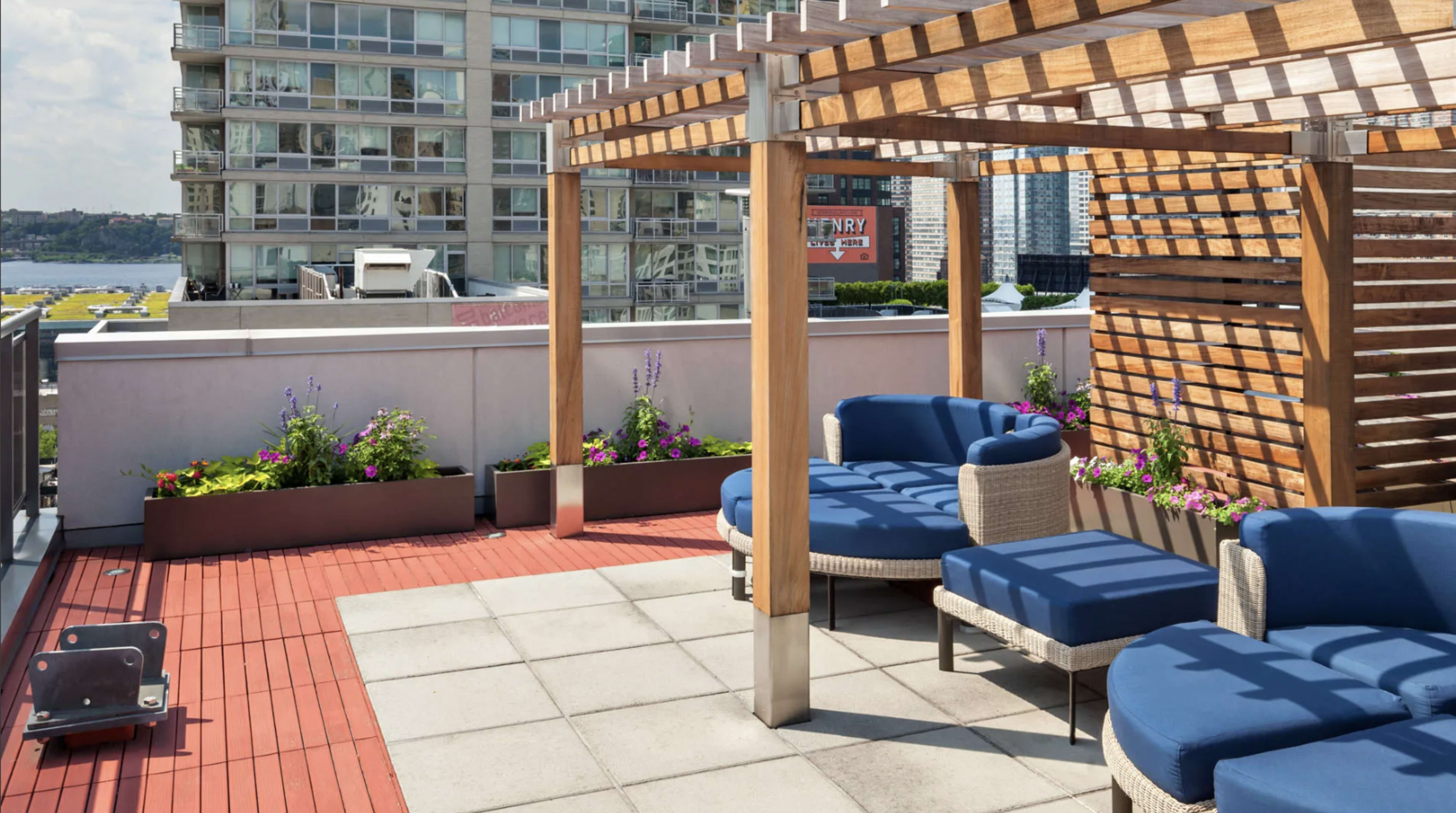 The image shows a rooftop terrace featuring wooden pergola structures, blue lounge chairs, and planters with colorful flowers, against a backdrop of a modern apartment building.