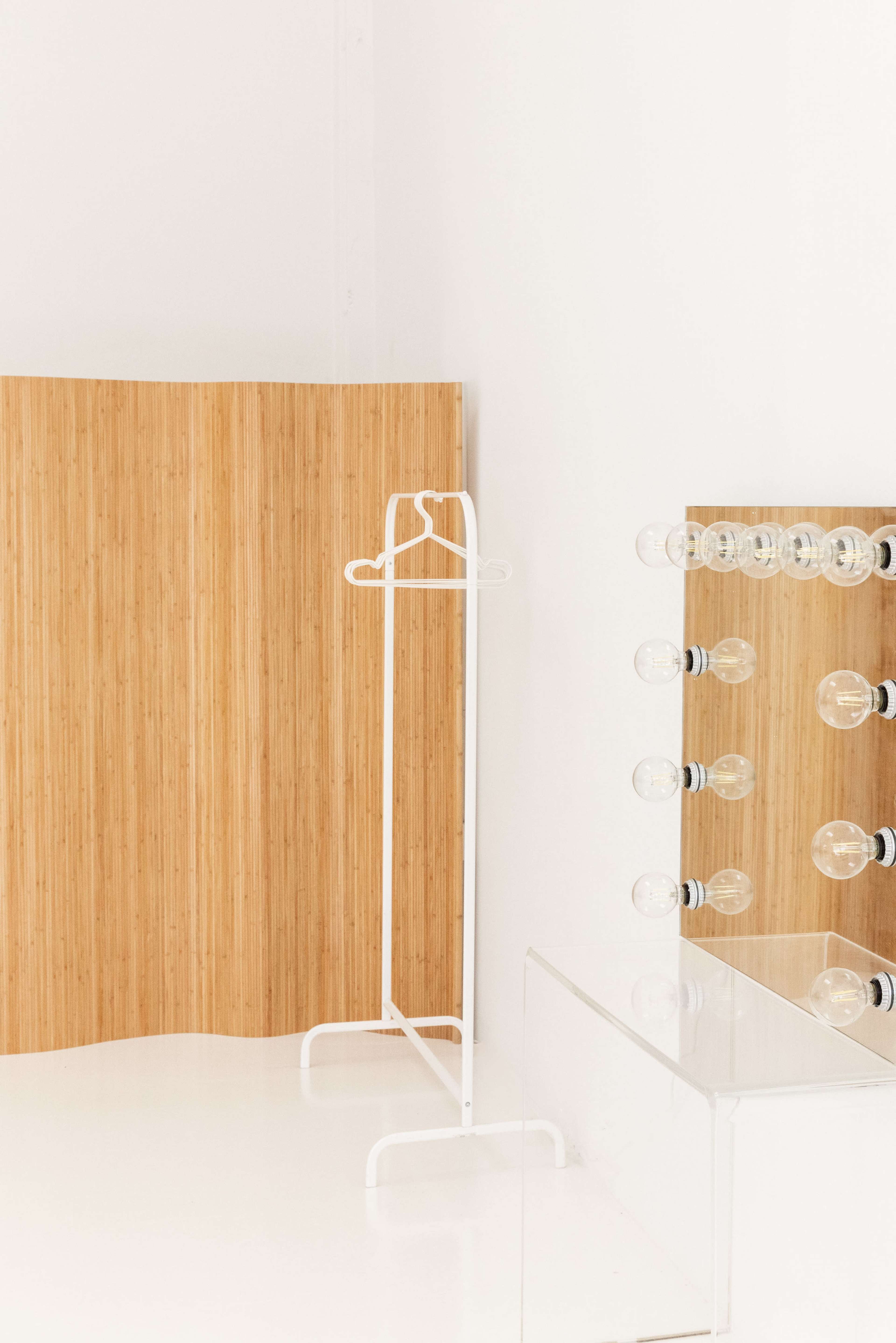 The image shows a minimalist dressing area featuring a wooden backdrop, a white clothing rack, and a mirror with illuminated bulbs.