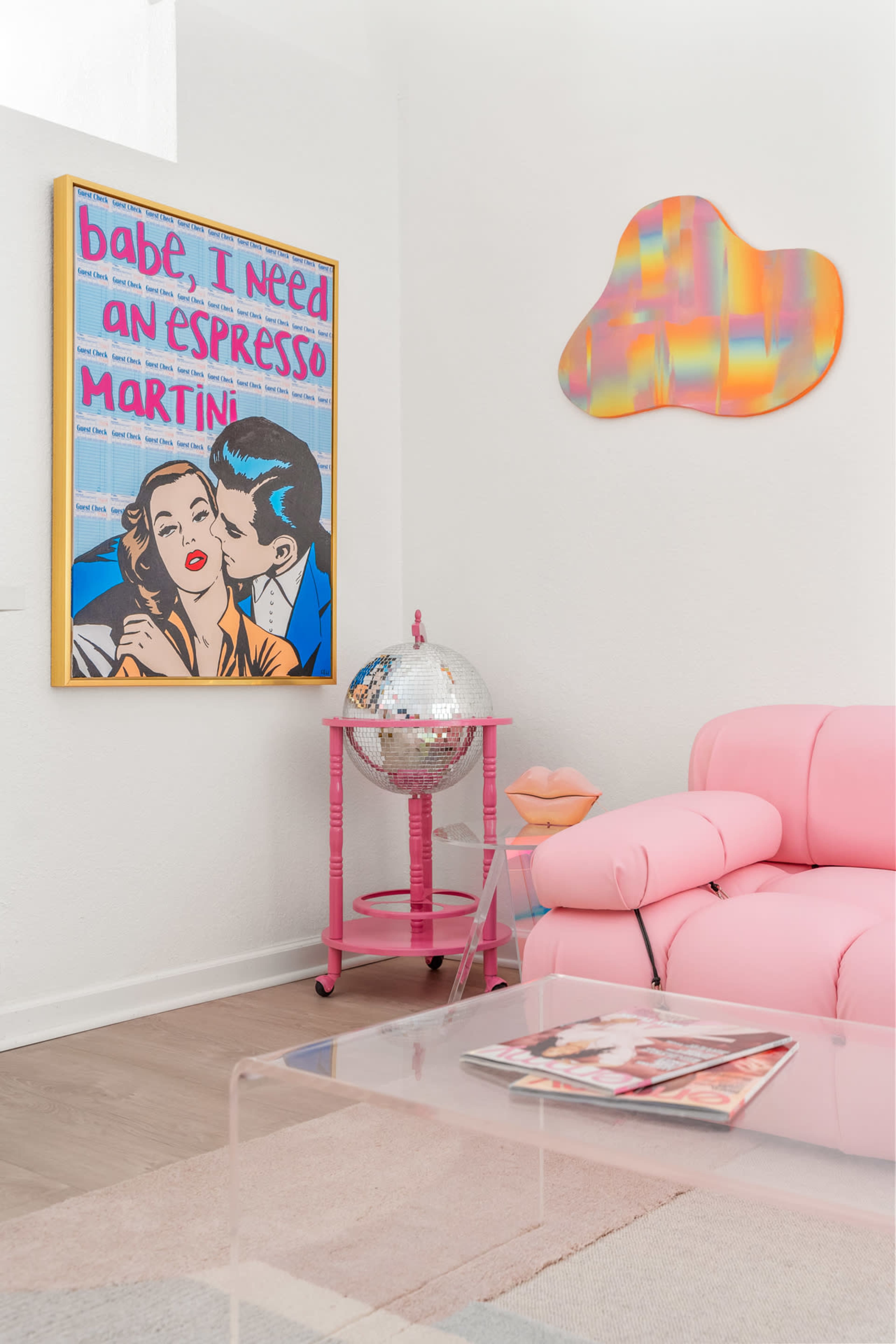 A pink sofa, a disco ball on a pink cart, and a framed pop art-style poster with a couple, set against a light-colored wall.
