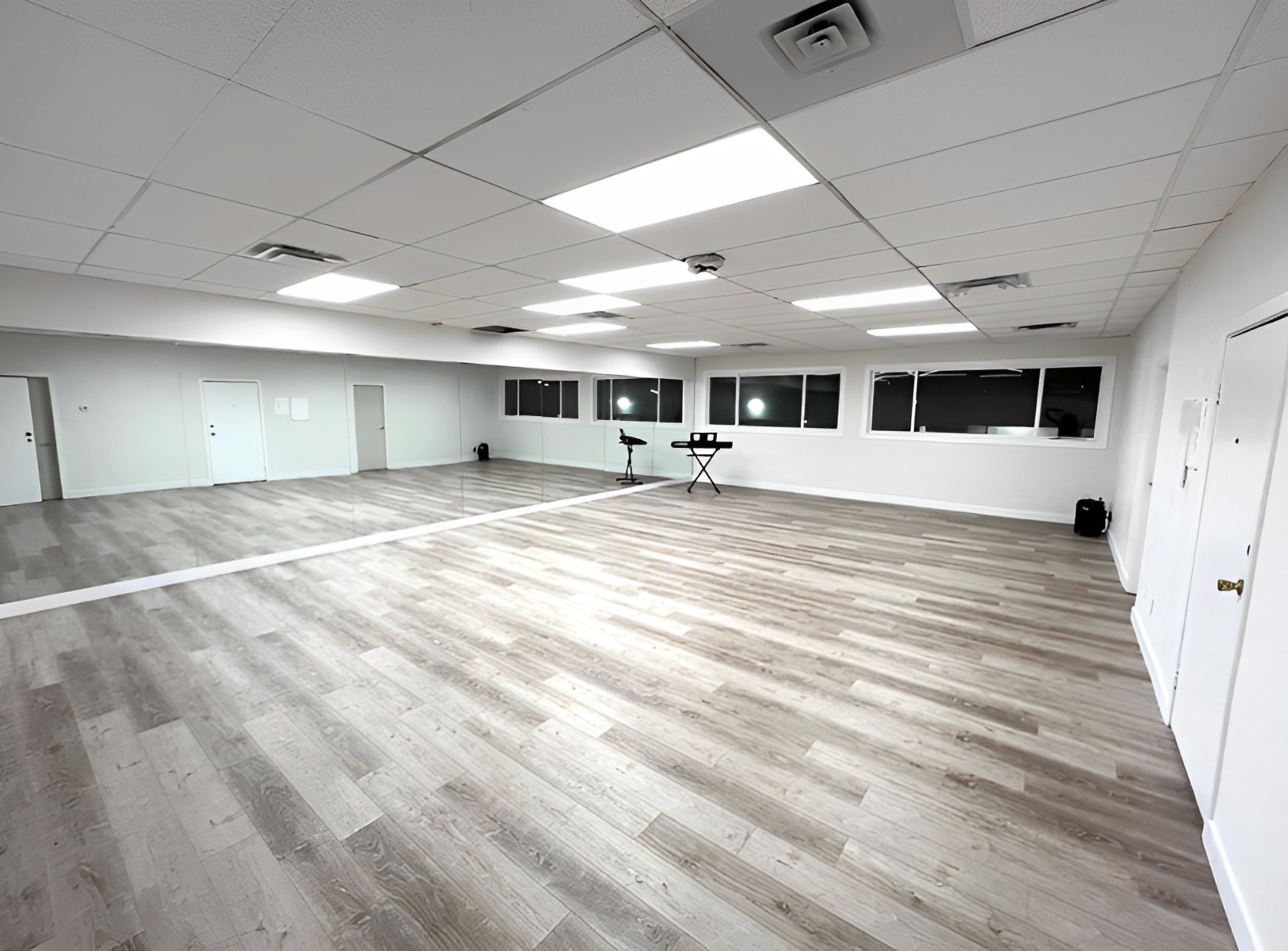 An empty dance studio features a large wooden floor, mirrors on one wall, and bright overhead lighting.