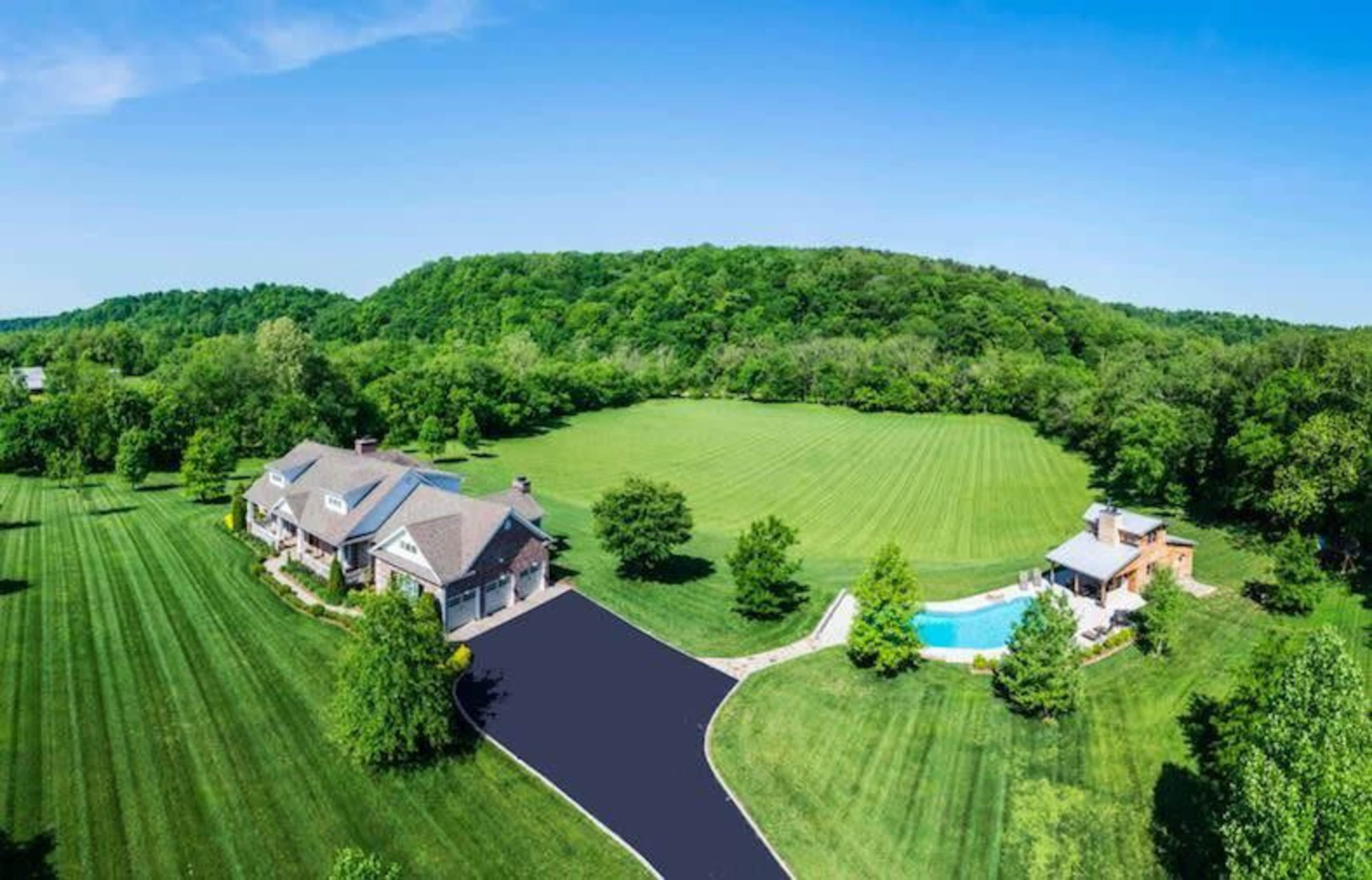 The image shows a large house with a pool surrounded by a manicured lawn and wooded hills in the background.