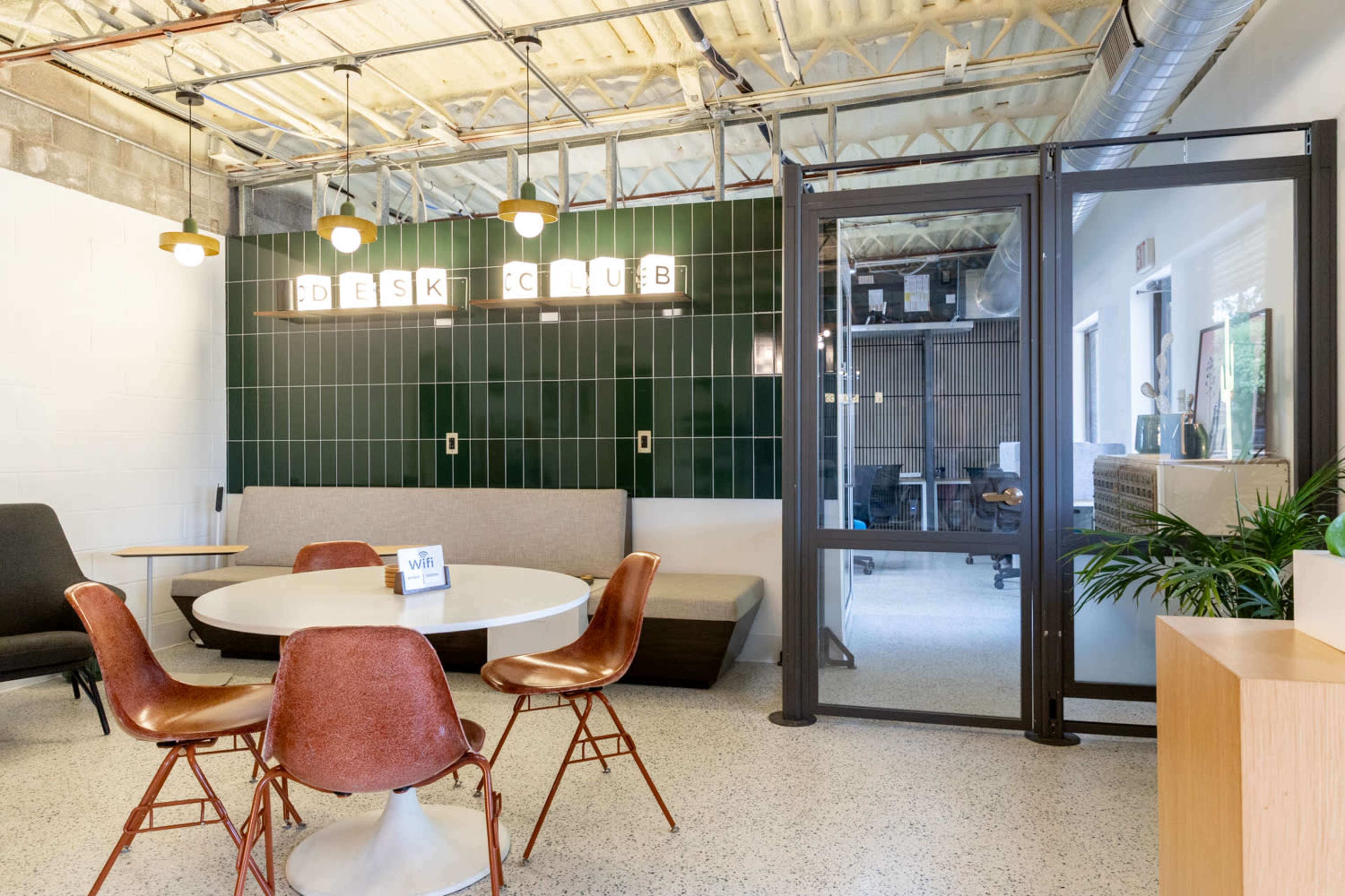 The image shows a modern coworking space with a round table, chairs, and a green tiled wall featuring the words "DESK CLUB" illuminated above a seating area.