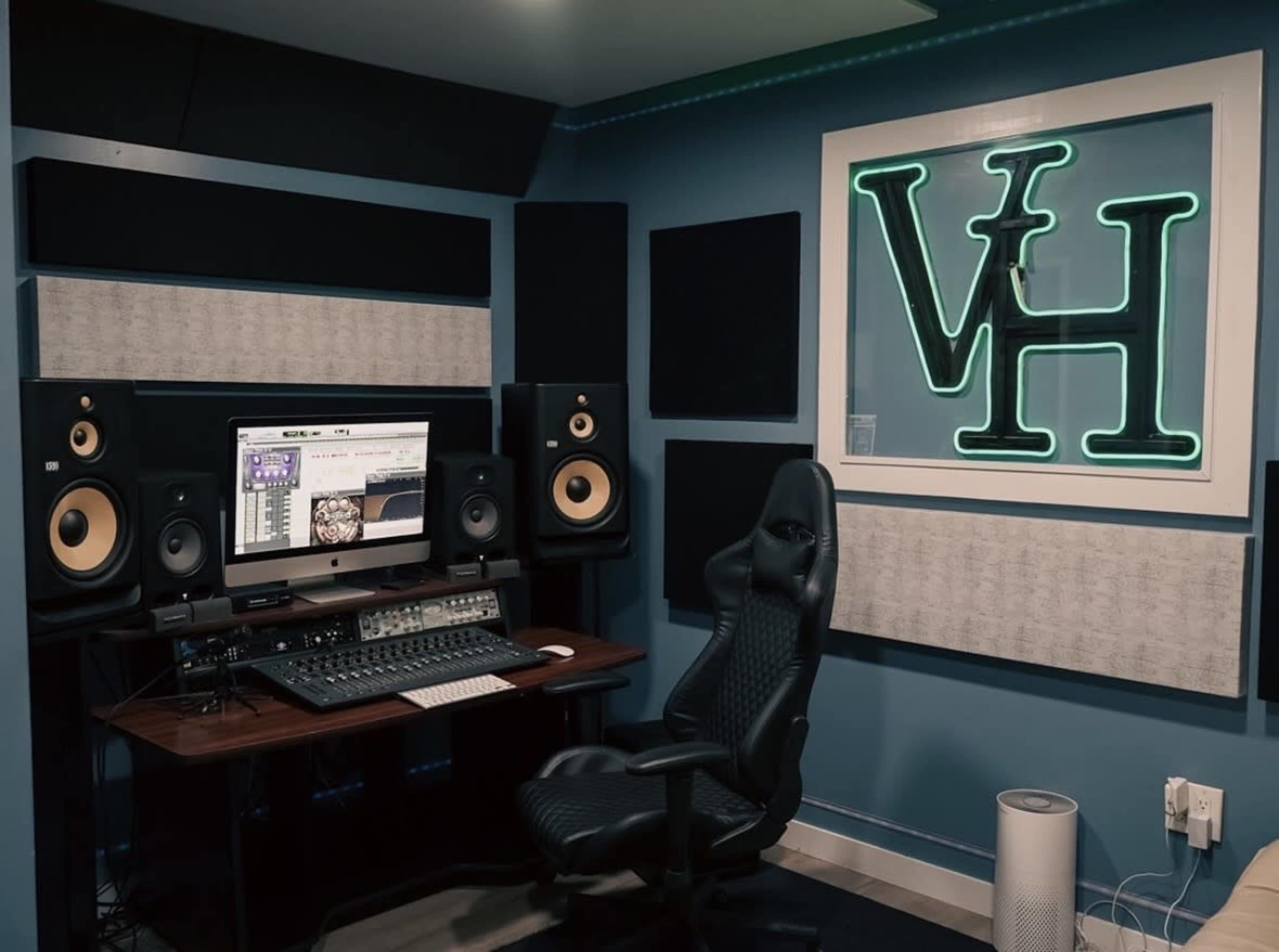 The image shows a music studio featuring a desk with computer equipment, studio monitors on either side, and a neon sign displaying "VH" on the wall.