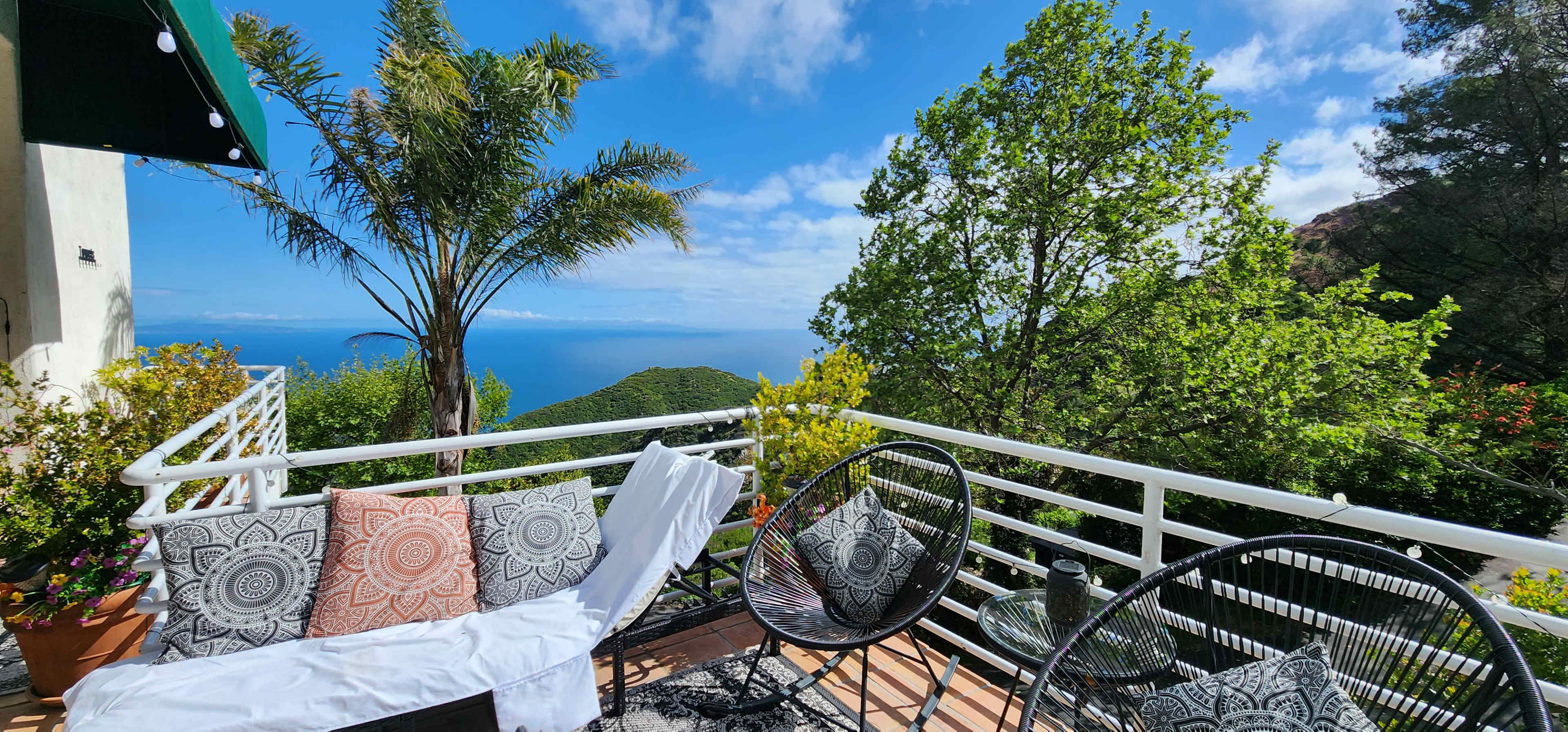 A balcony overlooks a coastal view surrounded by trees, featuring black wicker chairs and decorative pillows.
