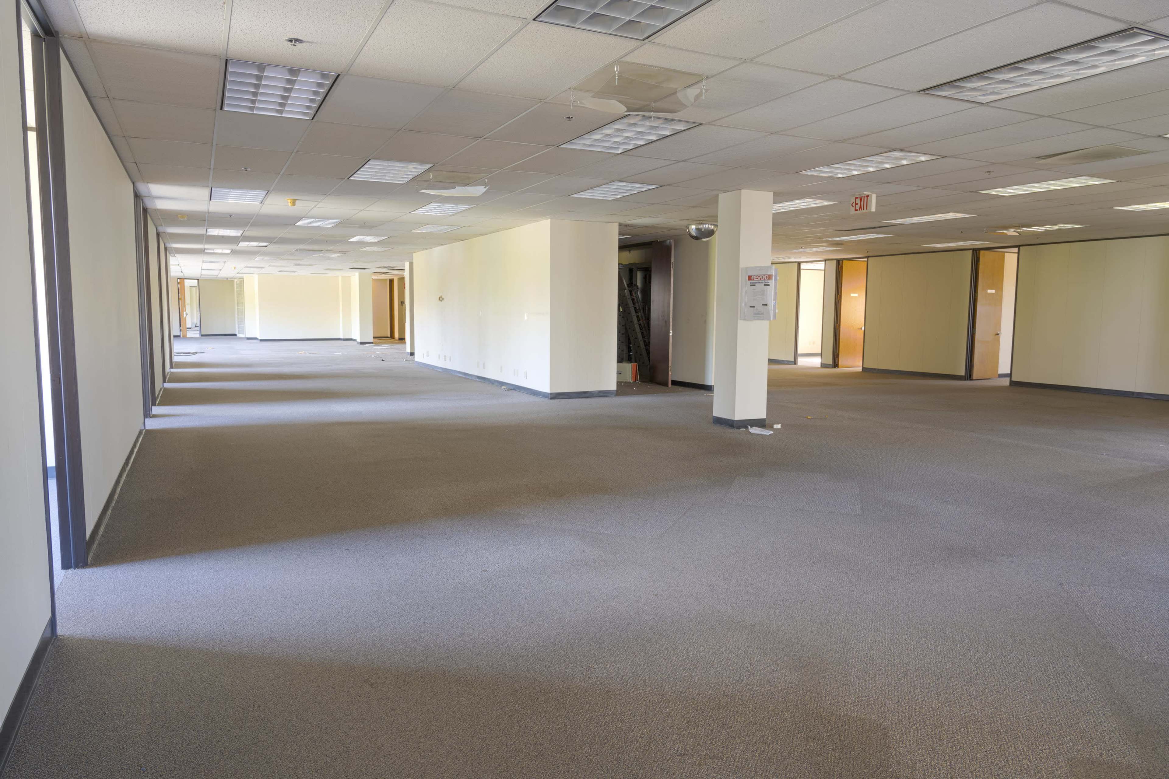 A vacant office space with gray carpet, beige walls, and empty rooms along either side.