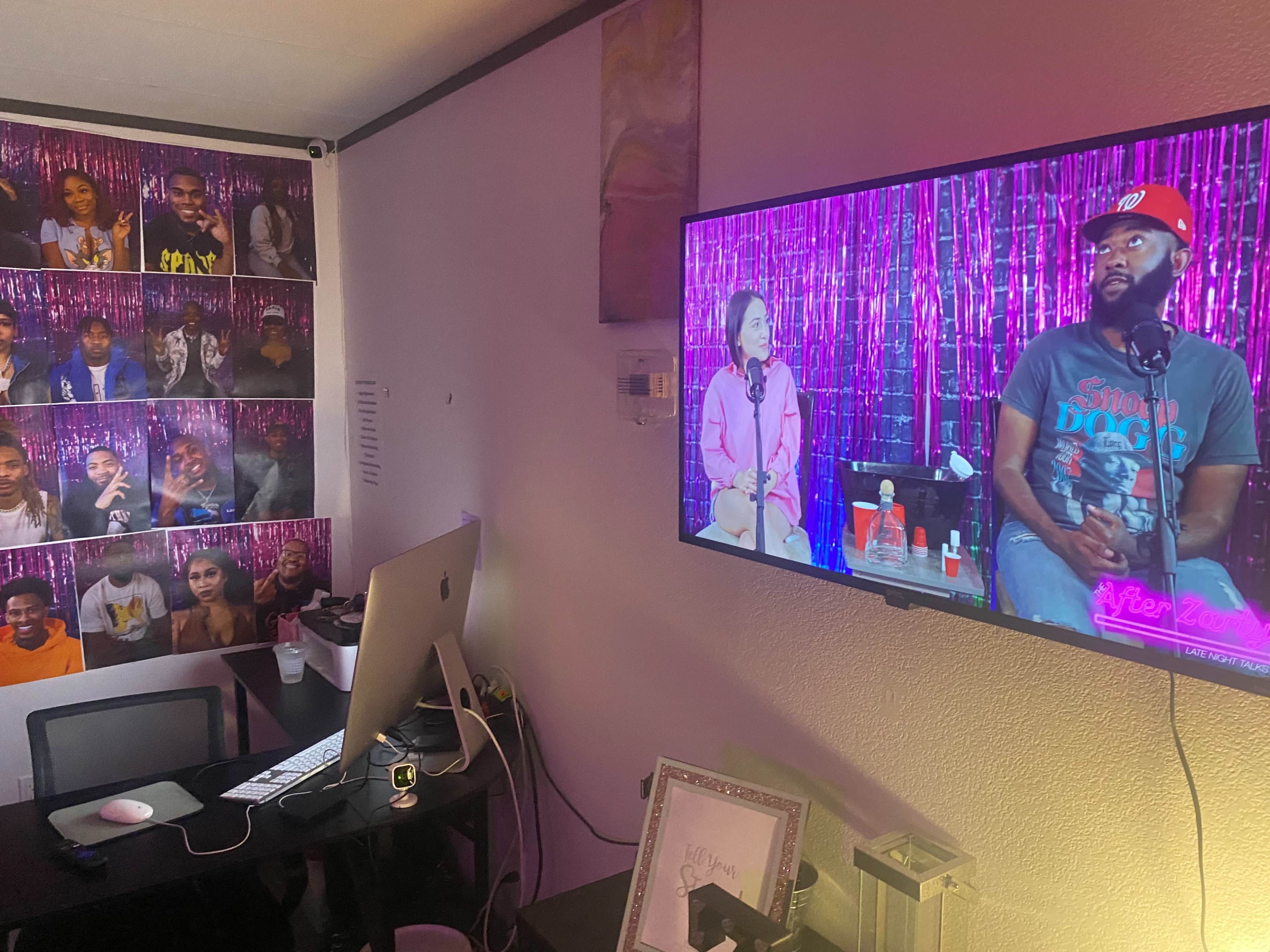 A television displays a talk show featuring two hosts, while the wall adjacent is covered with various photos of individuals.