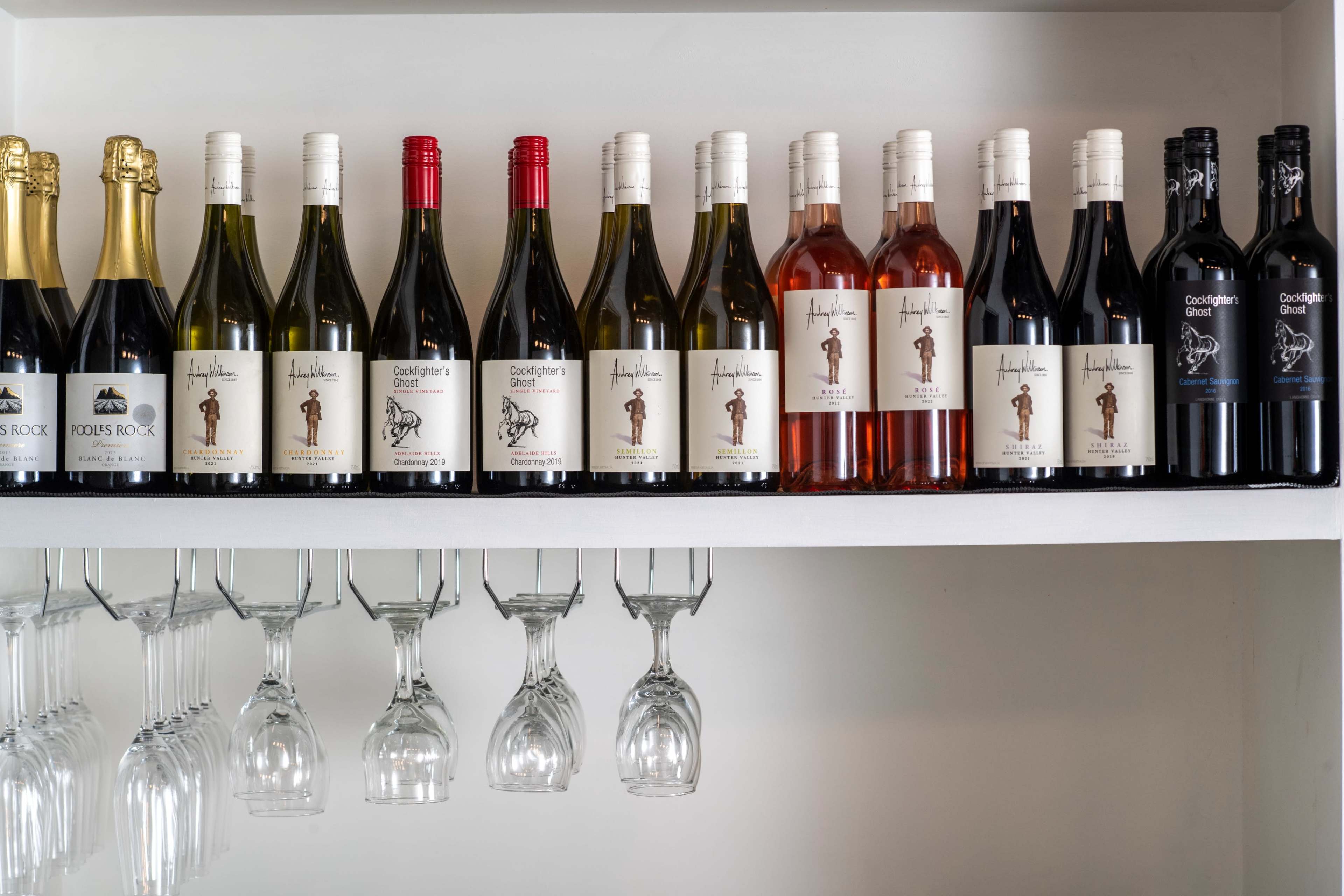 A shelf displays a variety of wine bottles in different colors and labels, alongside hanging wine glasses.
