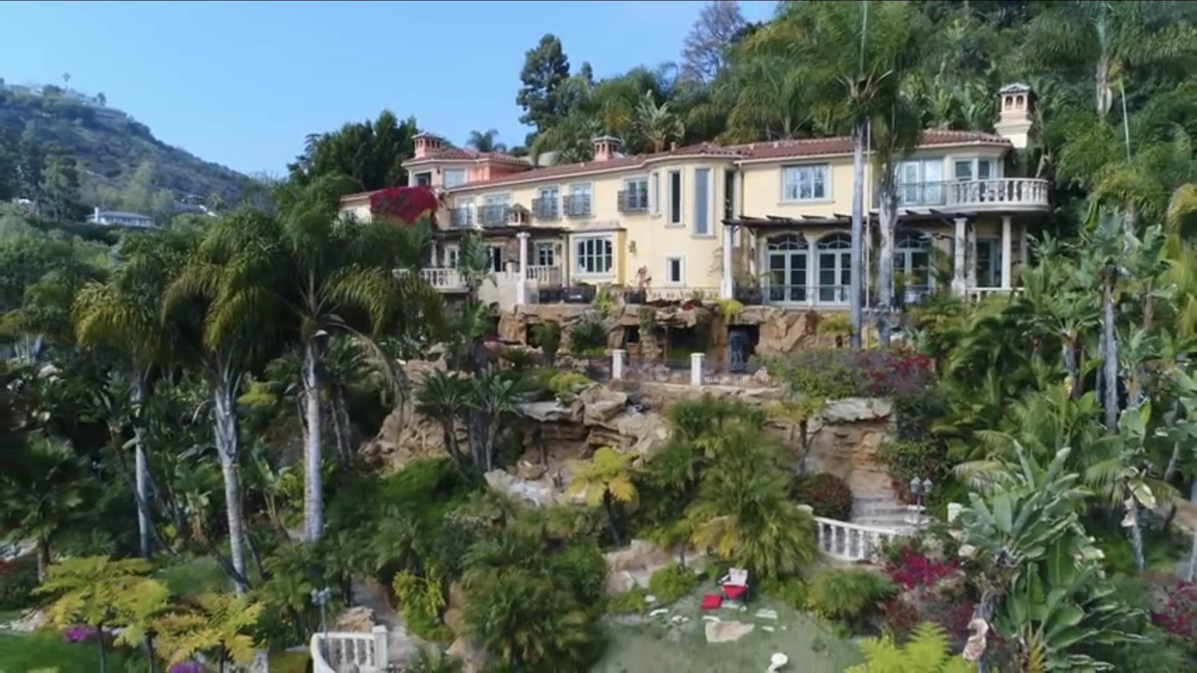 A large, multi-level mansion surrounded by lush greenery and colorful landscaping is situated on a hillside.