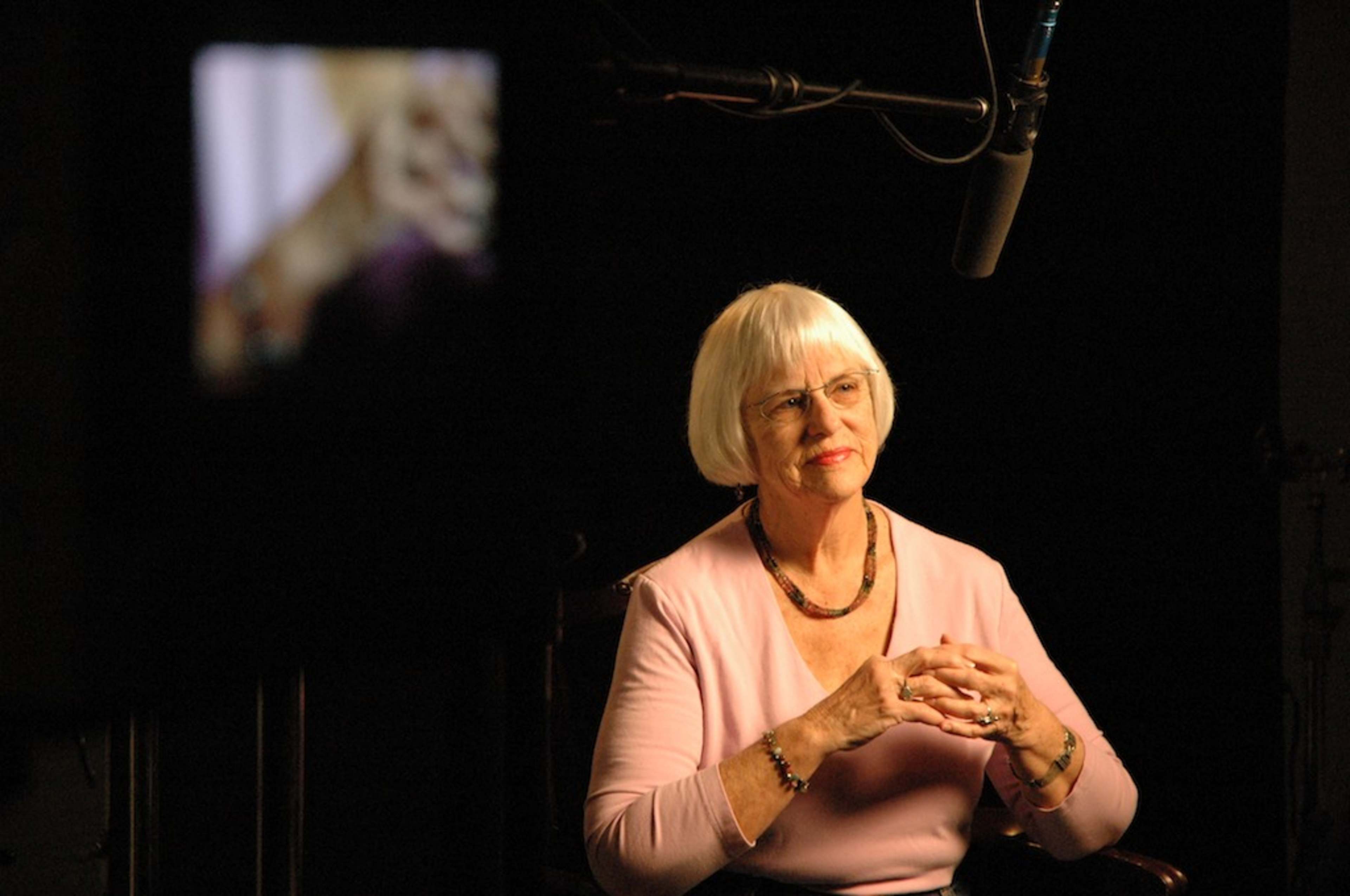 A woman with short, gray hair sits with her hands clasped in front of her, while a microphone and camera are positioned nearby.
