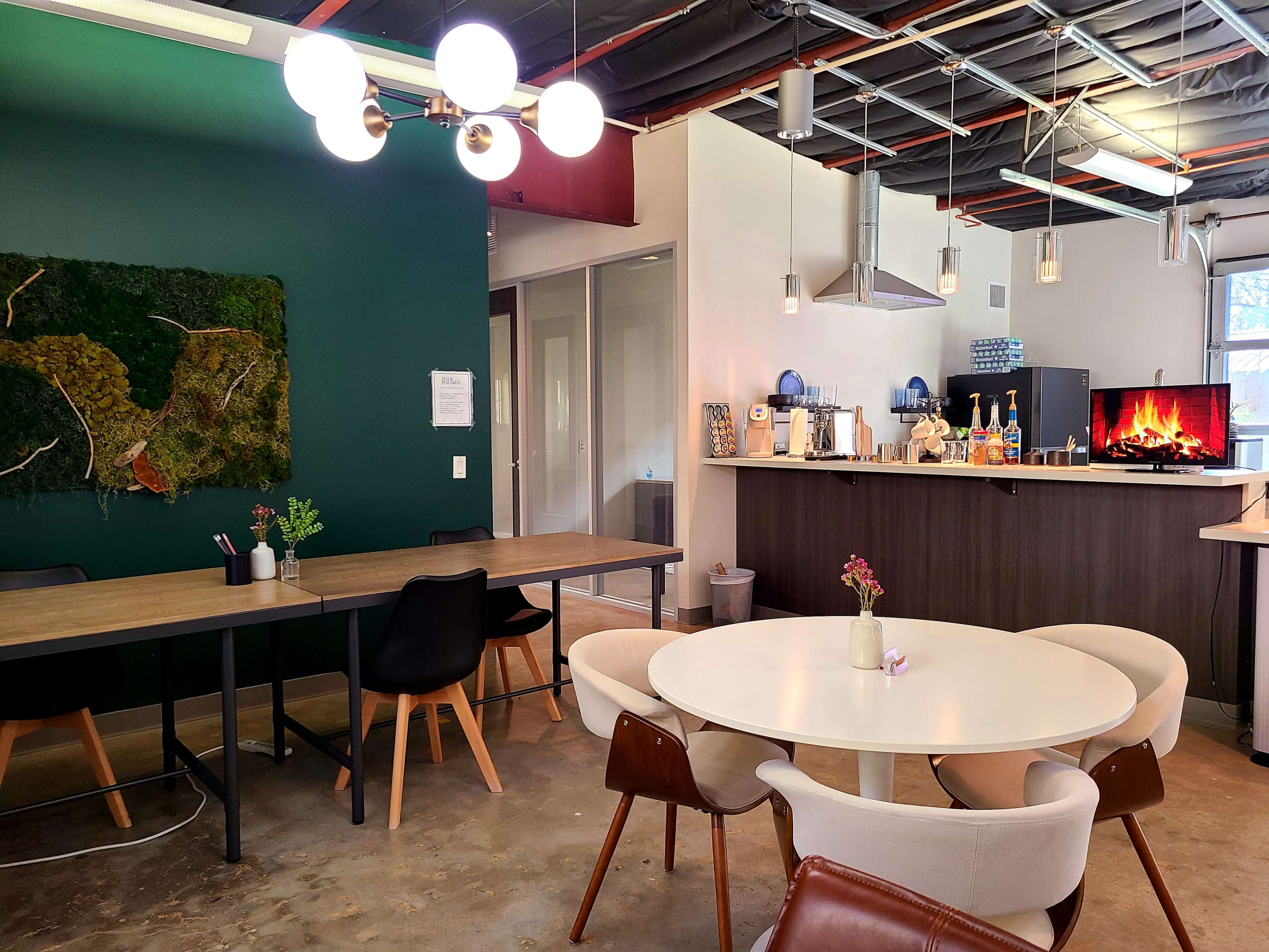 A modern communal kitchen and dining area with a mix of seating arrangements, including a round table and long table, accompanied by a green accent wall and a kitchen counter featuring various appliances.