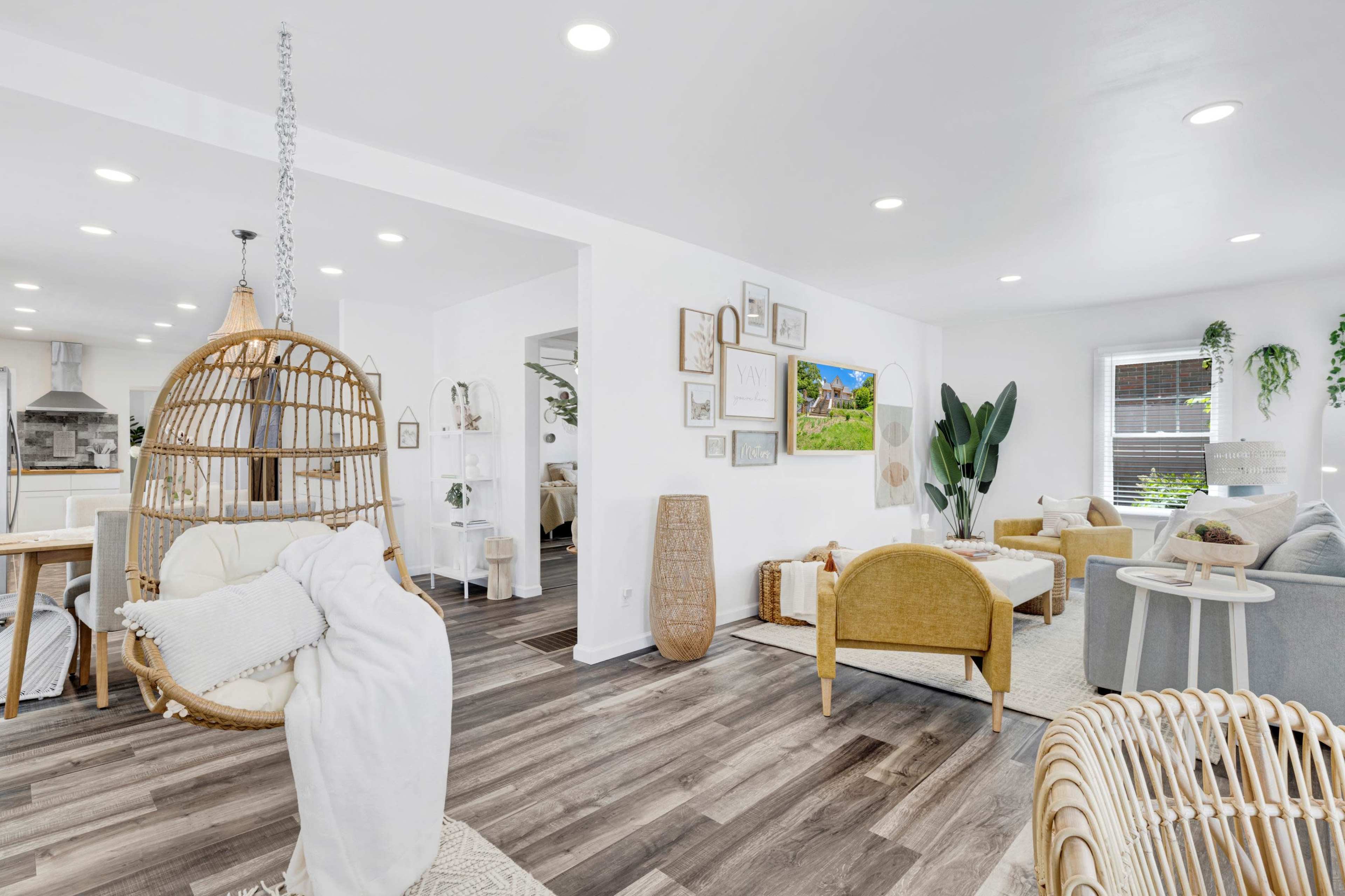 A modern living area features a hanging wicker chair, two couches, and a decorative wall adorned with framed artwork and indoor plants.