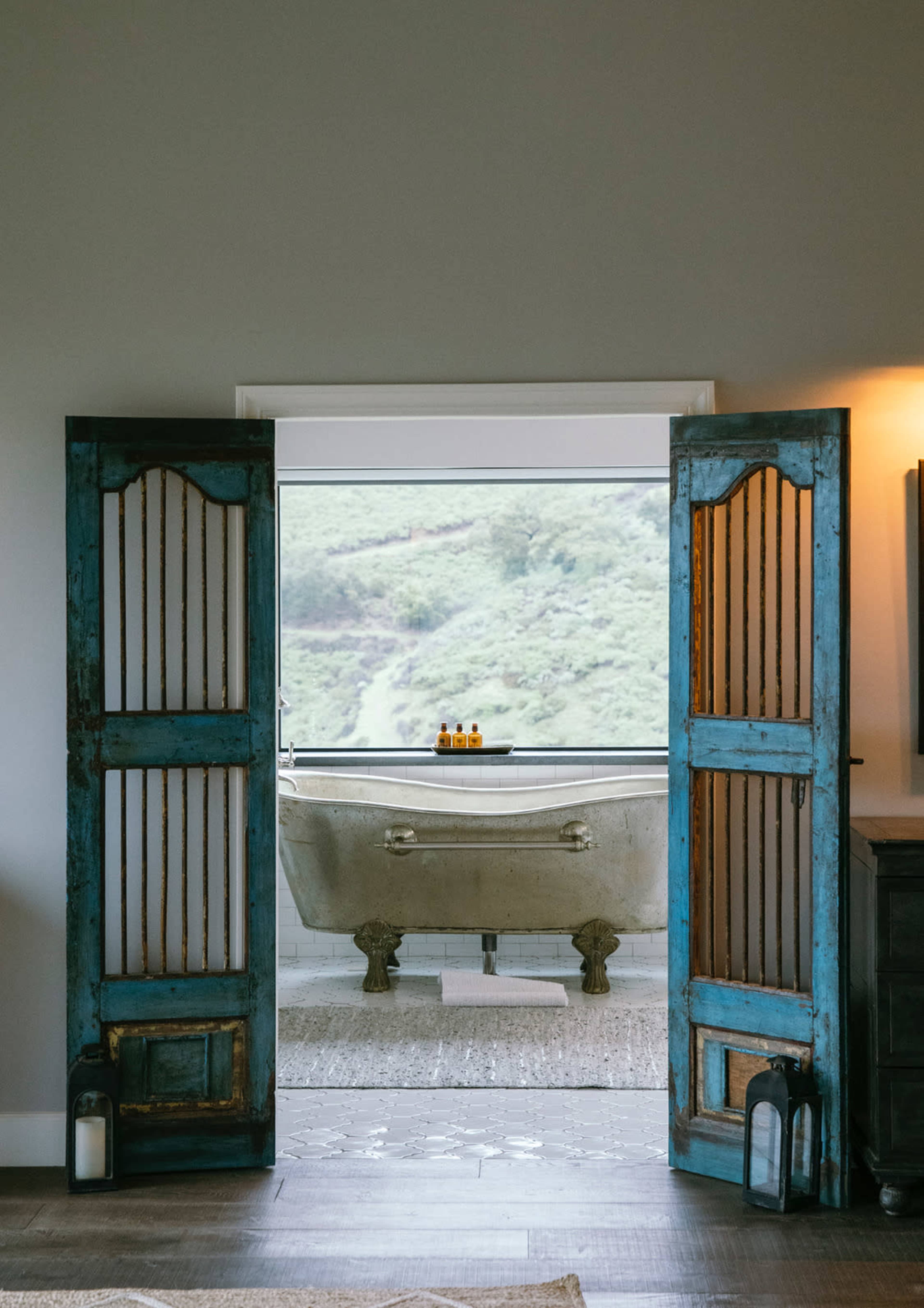 An antique bathtub is positioned in front of an open window, framed by blue wooden doors, revealing a scenic view of green hills.