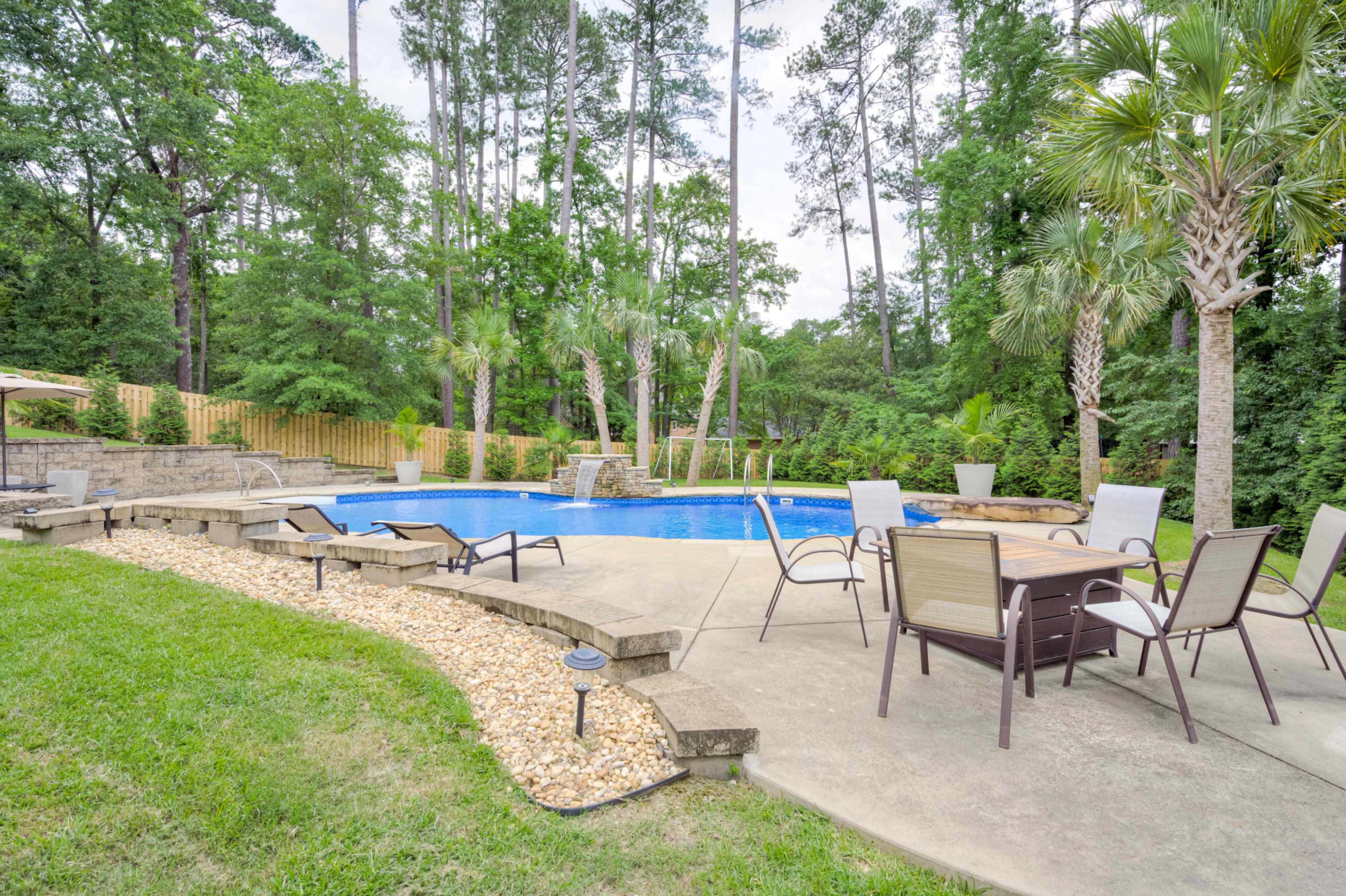 The image shows a residential backyard featuring a swimming pool surrounded by greenery, stone pavers, and outdoor seating.