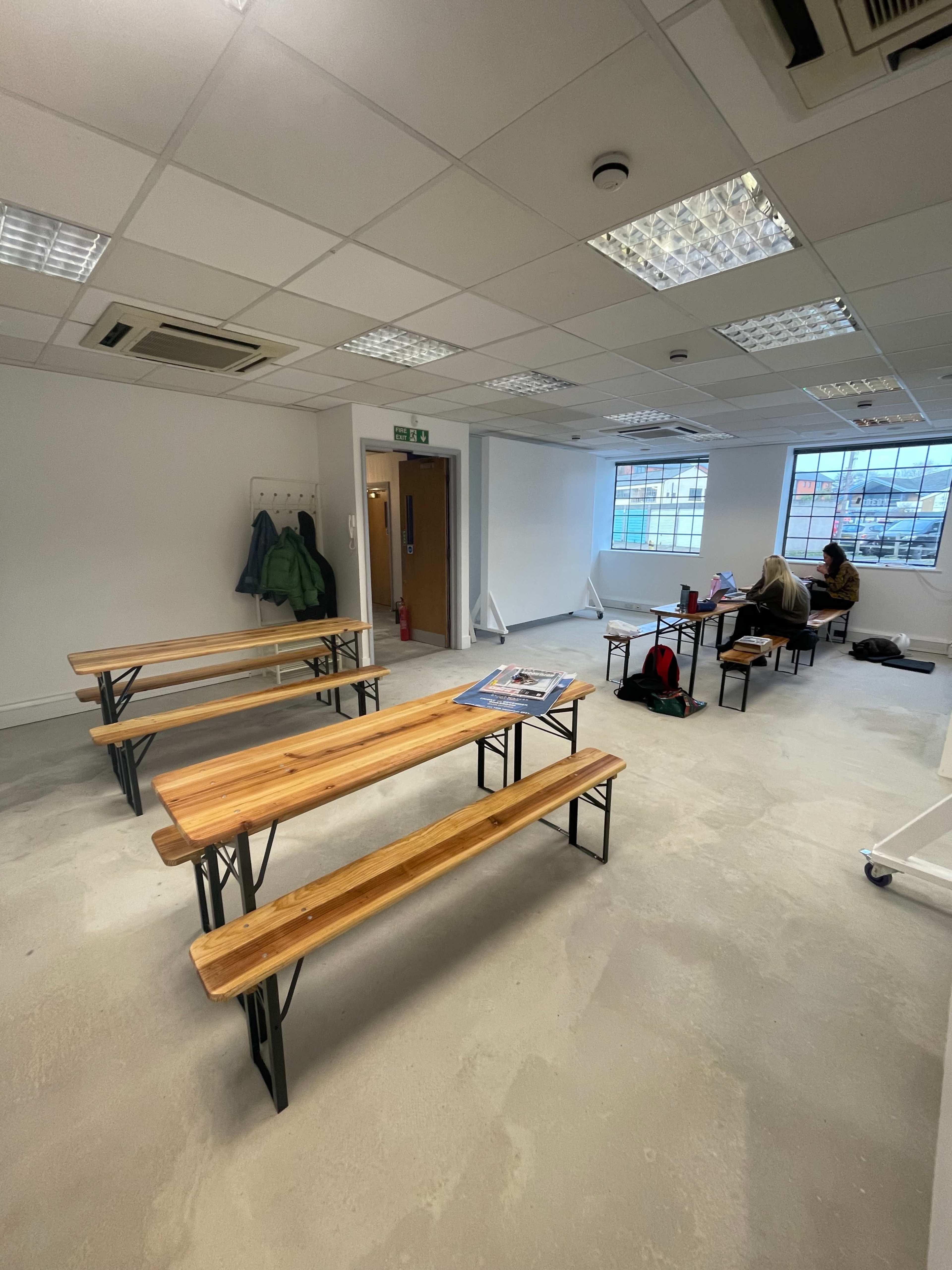 A nearly empty room with several wooden benches, a few scattered bags, and a person sitting at a table near a large window.