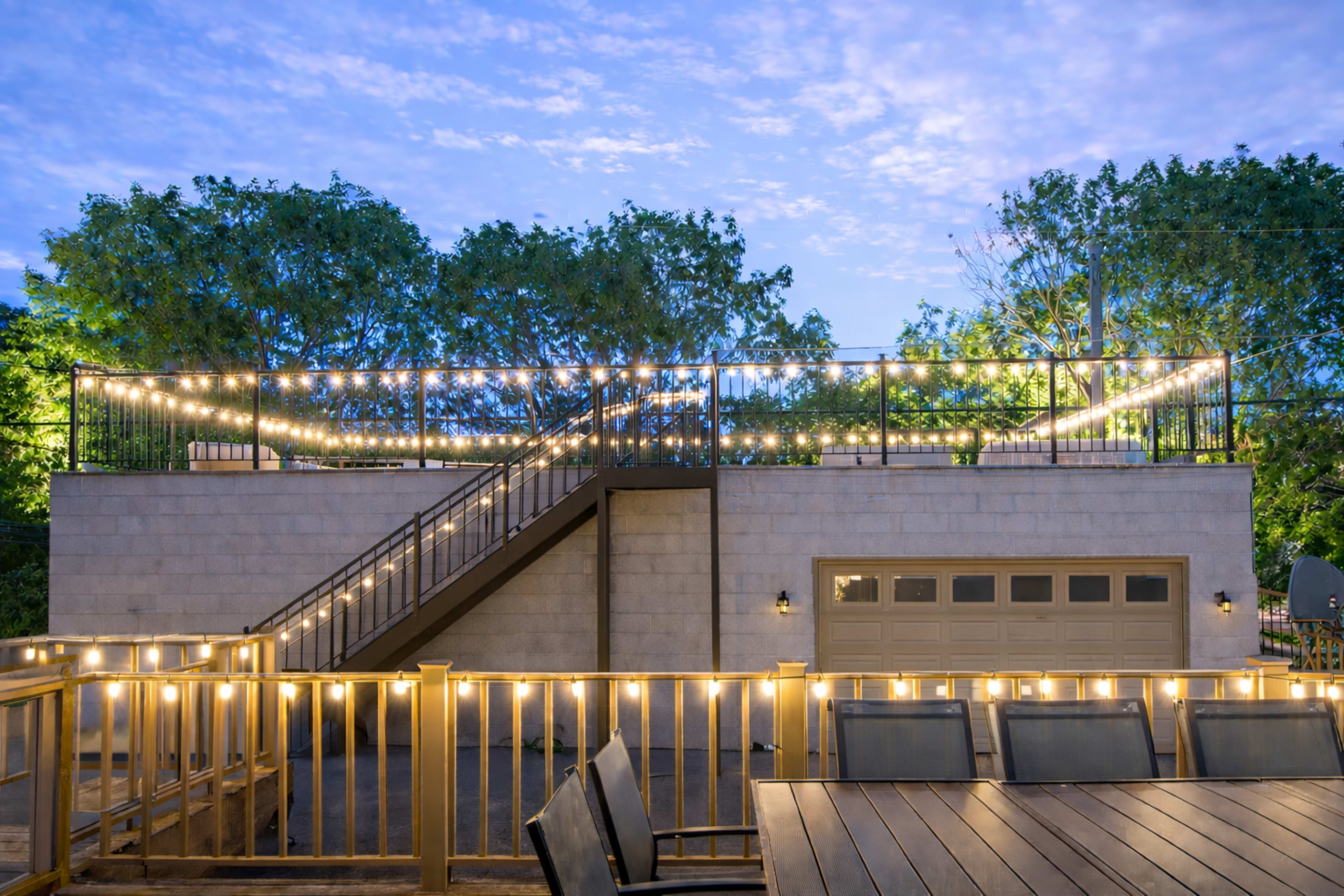 A rooftop space with string lights and a staircase overlooks a garage, surrounded by trees under a cloudy sky.