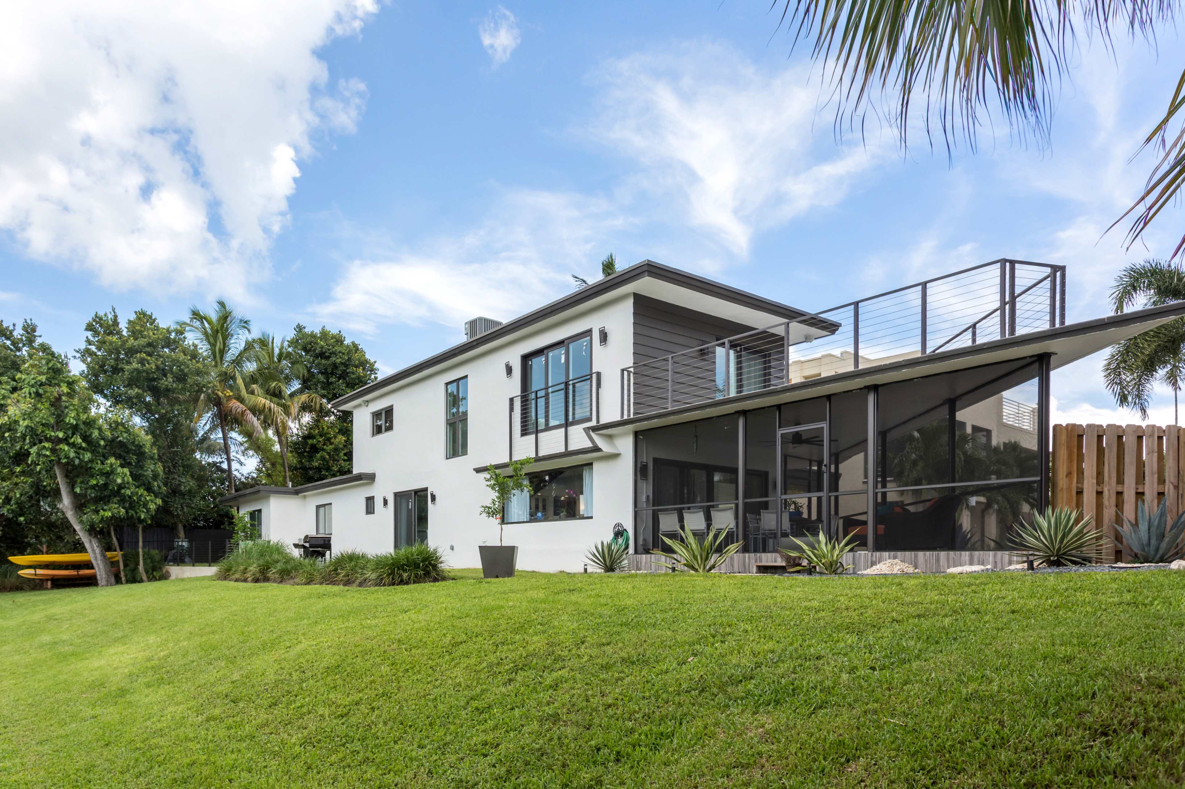 A modern two-story house with a deck overlooks a neatly manicured lawn and palm trees.