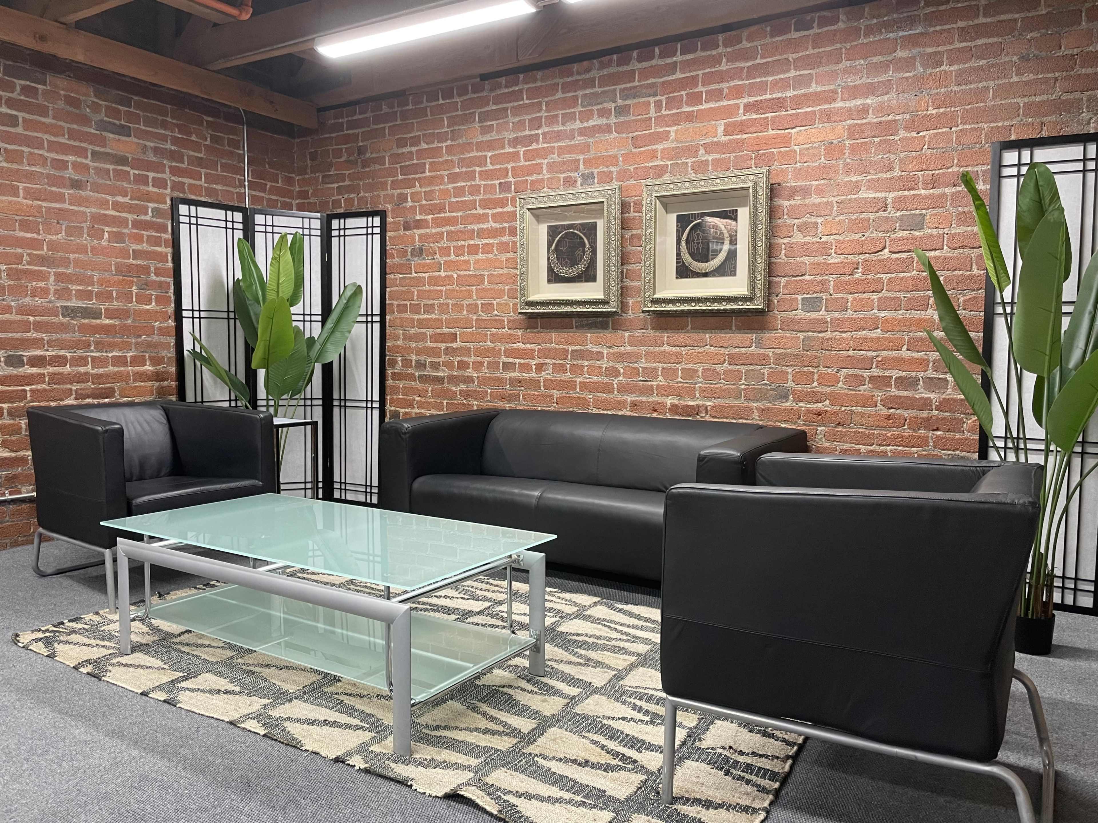 A seating area features two black leather sofas, a glass coffee table, and potted plants, set against a brick wall with framed artwork.