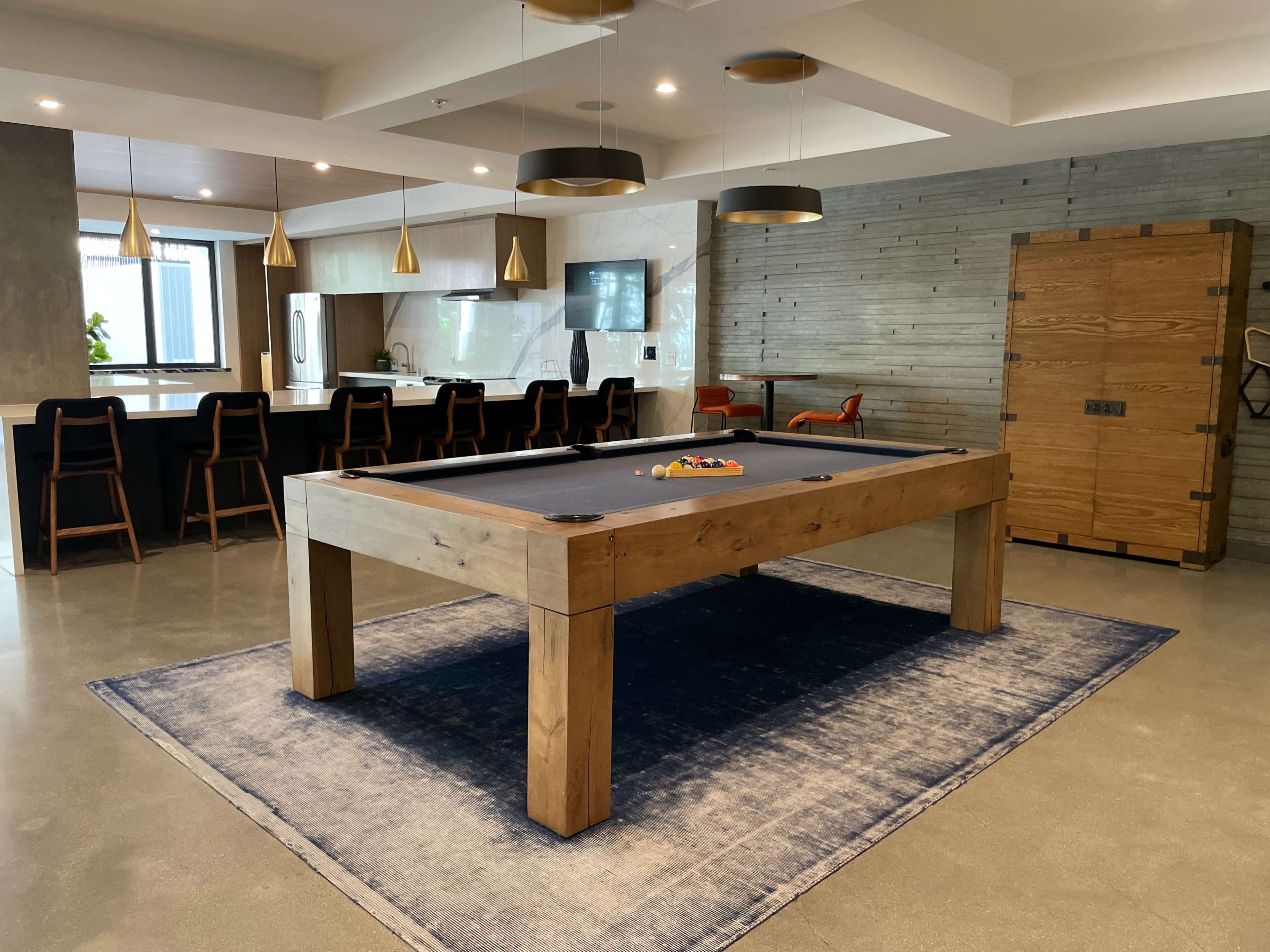 A spacious recreation room featuring a wooden billiards table on a blue rug, surrounded by modern bar seating and a stylish kitchen area.