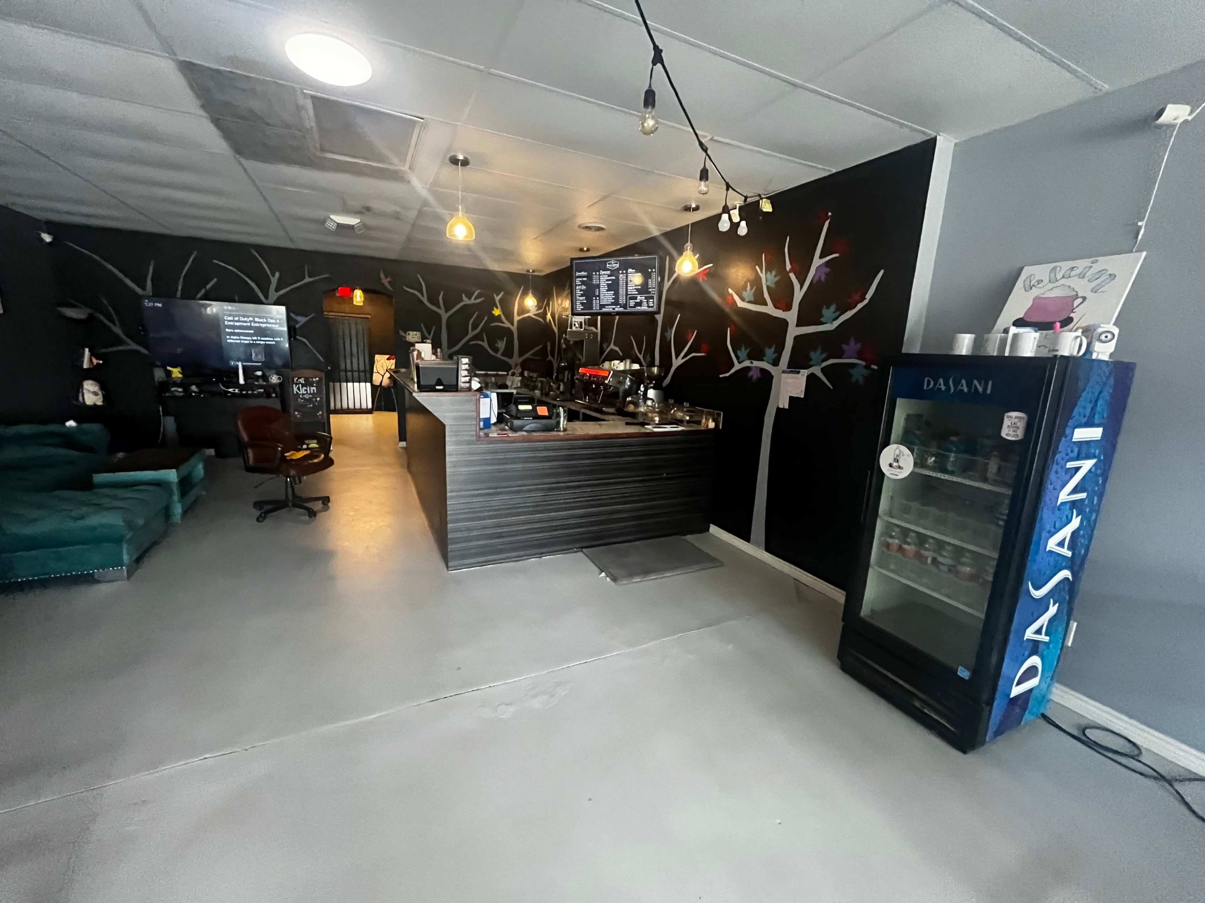 A modern cafe interior with a bar counter, seating area, and a refrigerator displaying bottled beverages against a wall adorned with white tree designs.
