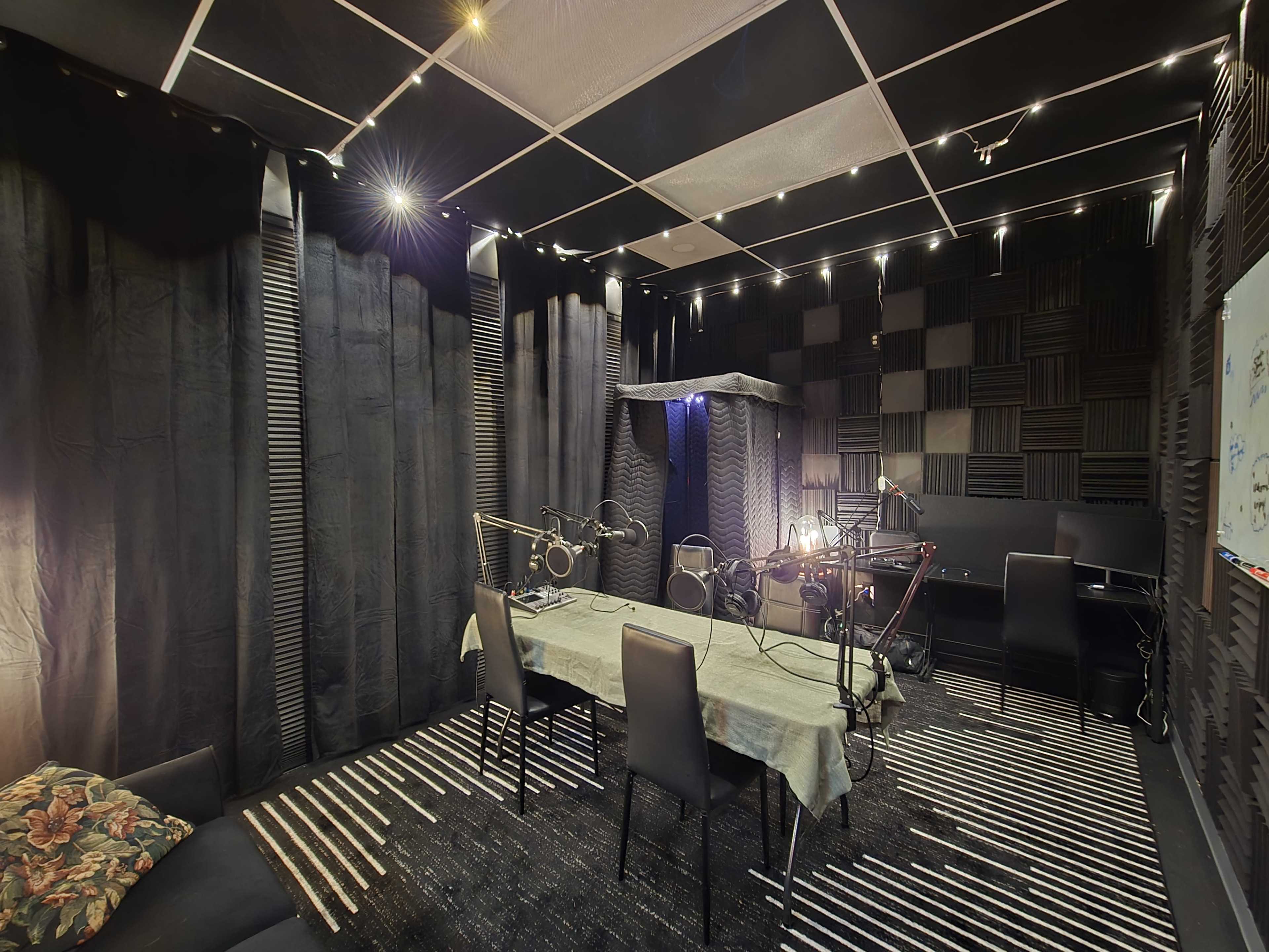 The image shows a soundproof recording studio with black walls, a table, several microphones, and chairs arranged within a space that features acoustic treatment.