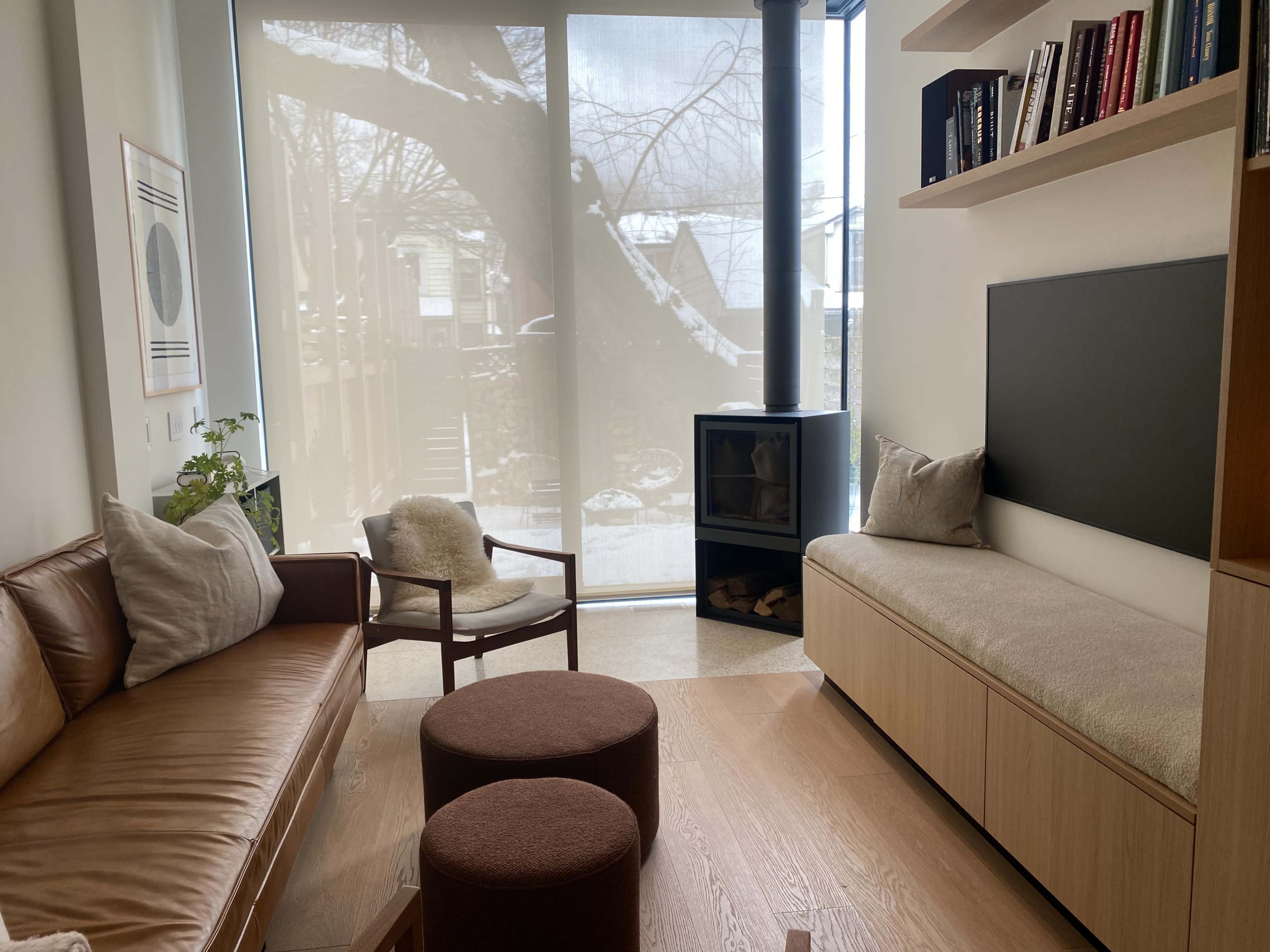 A modern living room with a leather sofa, two round stools, a wooden bench with a cushioned top, and a large window covered by sheer curtains that reveal a snowy outdoor scene.