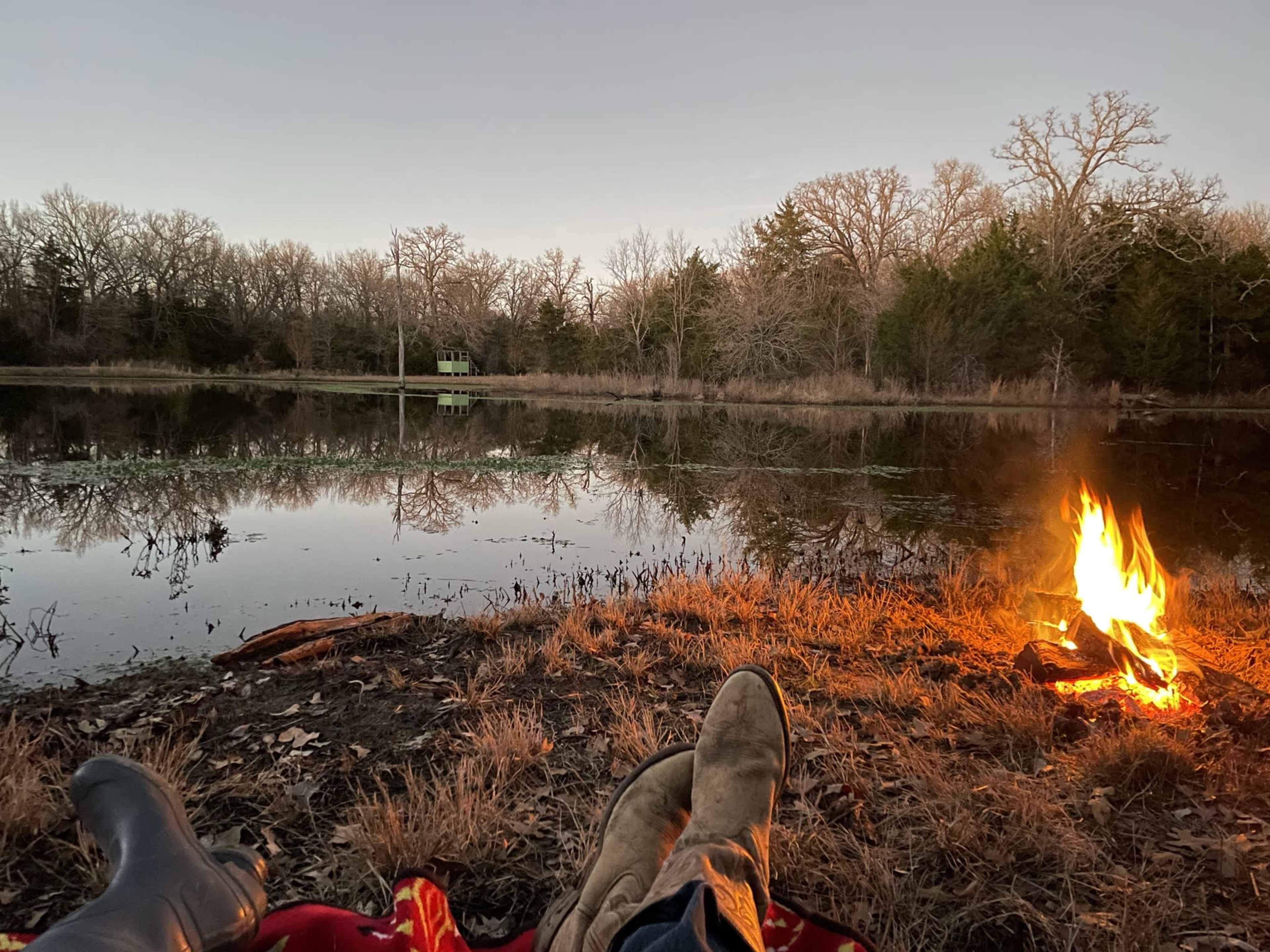 The scene shows a pair of boots resting on the ground beside a fire and a calm pond, with trees reflecting in the water under a twilight sky.