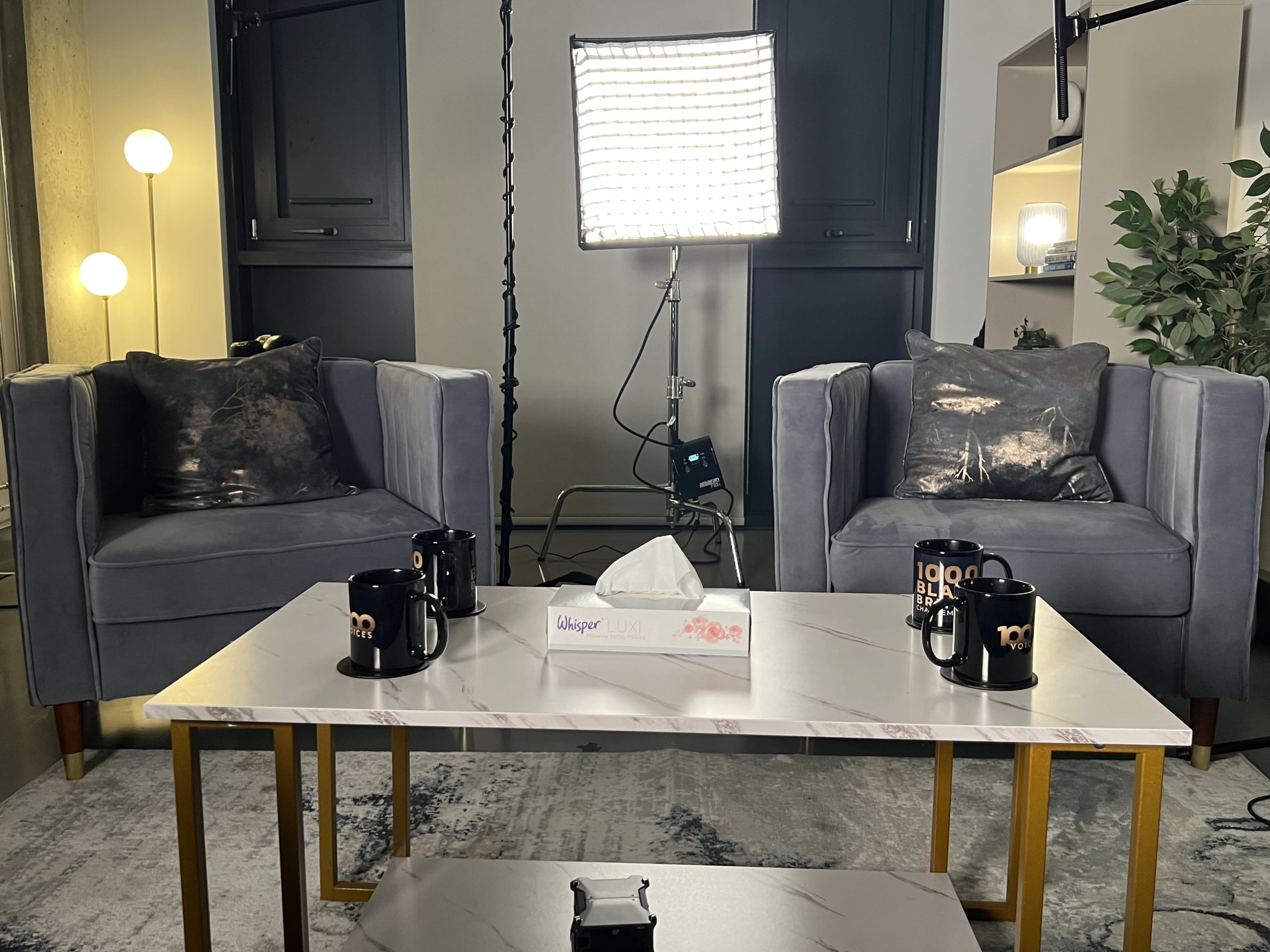 A cozy interview setup with two gray armchairs, a marble coffee table, a tissue box, and mugs placed on the table, illuminated by a bright studio light.
