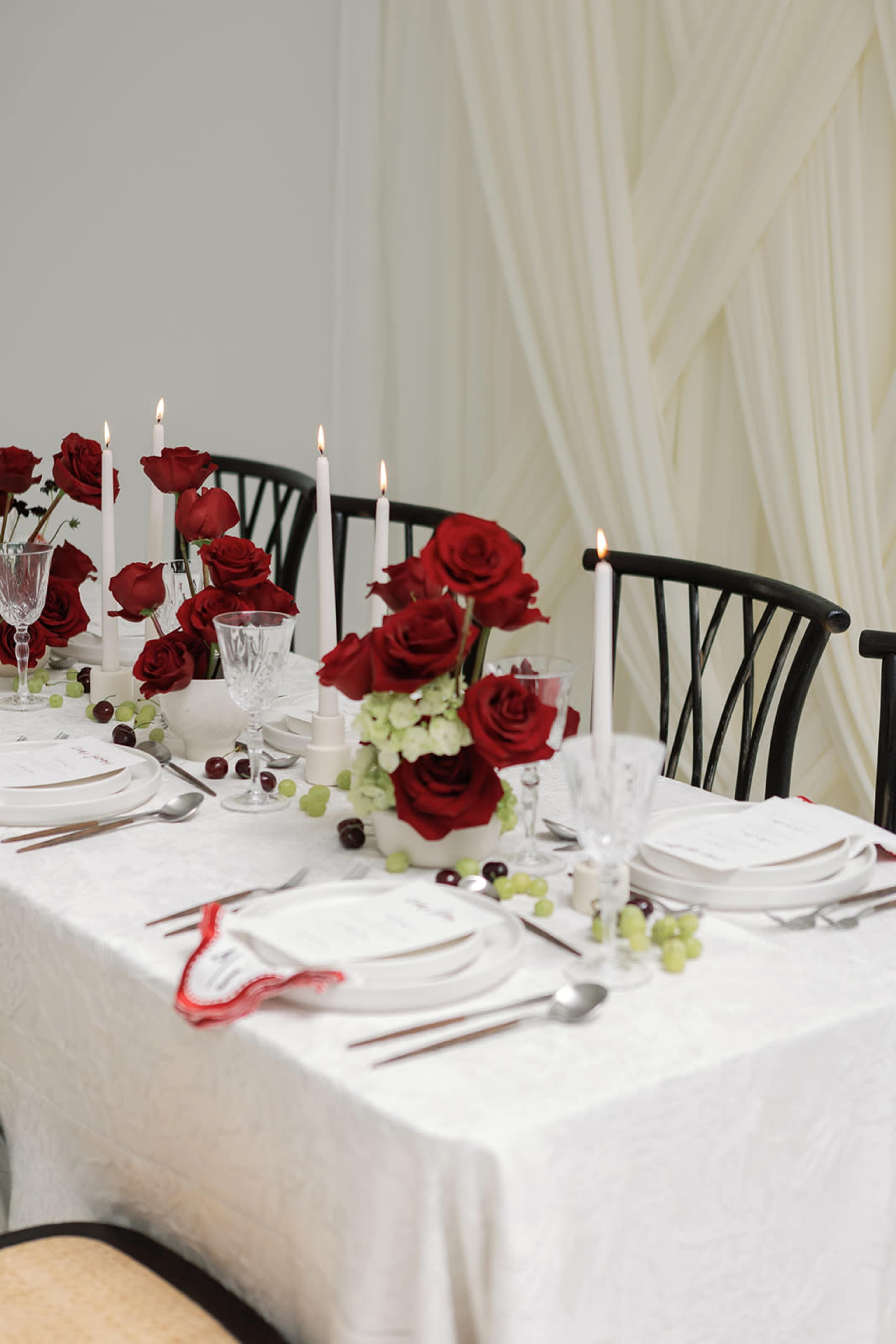 The image shows a elegantly set dining table with white tablecloth, adorned with red roses, candles, and glassware, complemented by fruit and decorative elements.