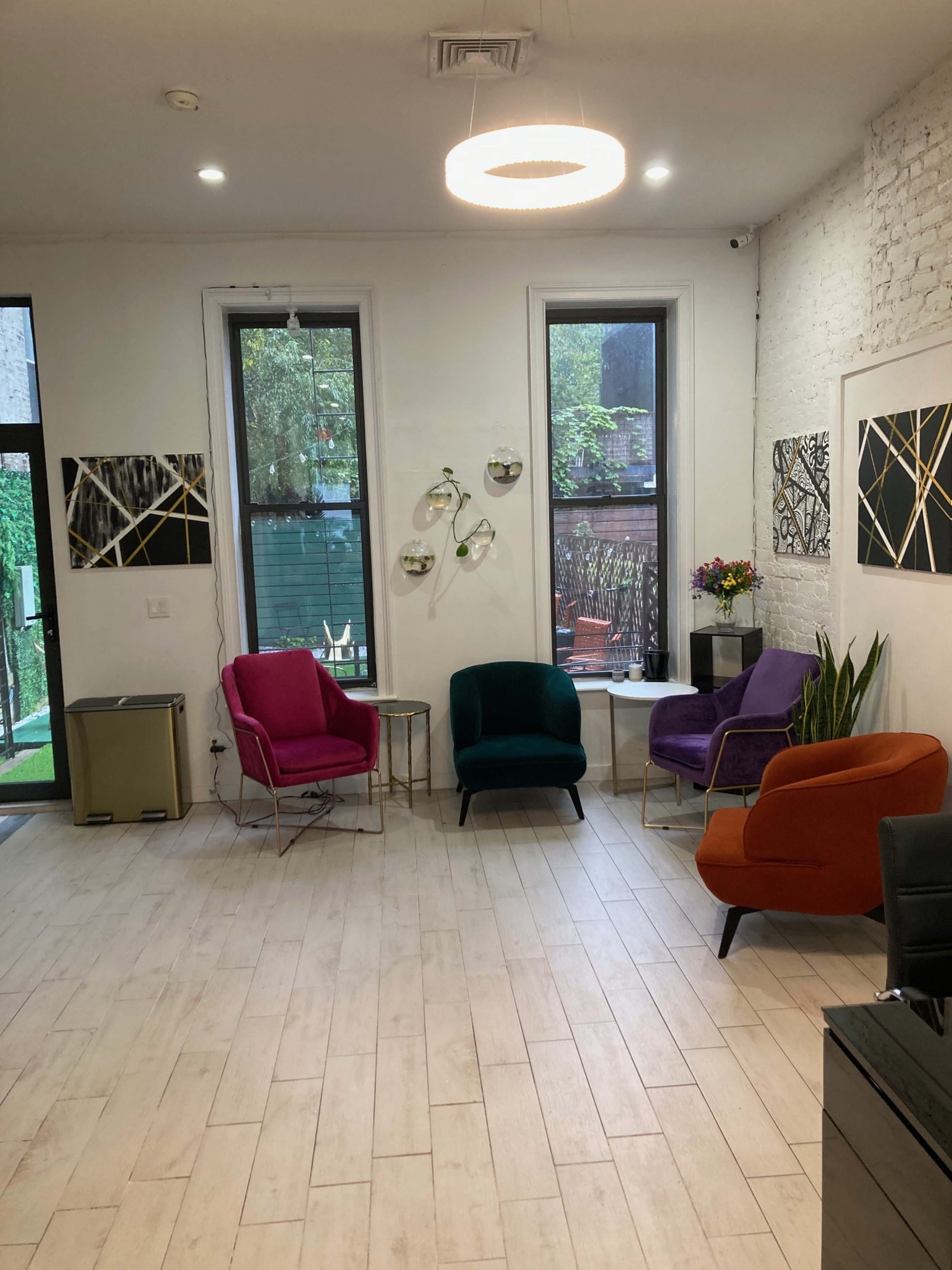 The image shows a modern interior space with four colorful chairs arranged around a small table, featuring large windows and decorative wall art.