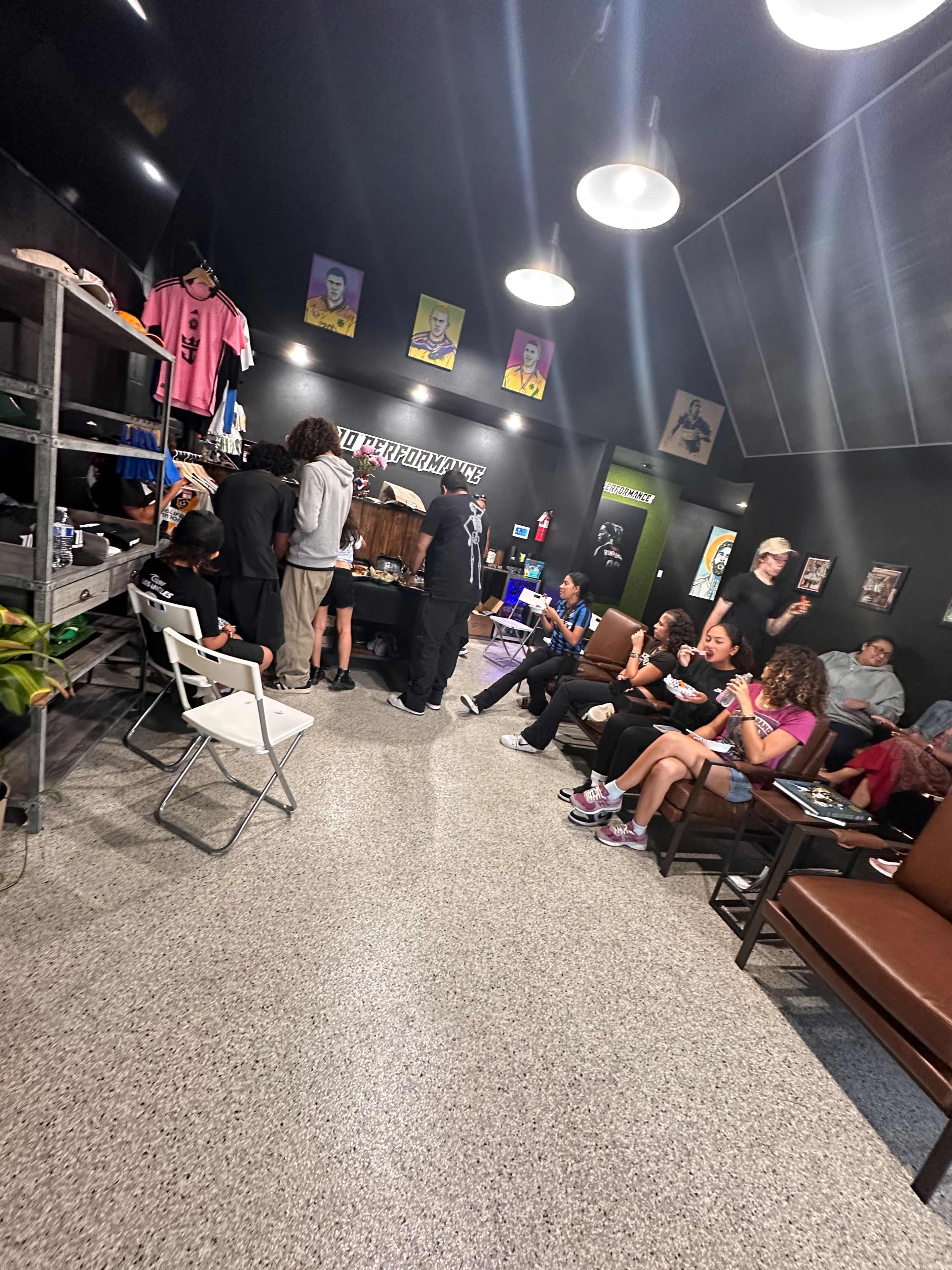 A group of people is gathered inside a shop with a modern interior, where some are seated while others are engaged in conversation or working at a counter.