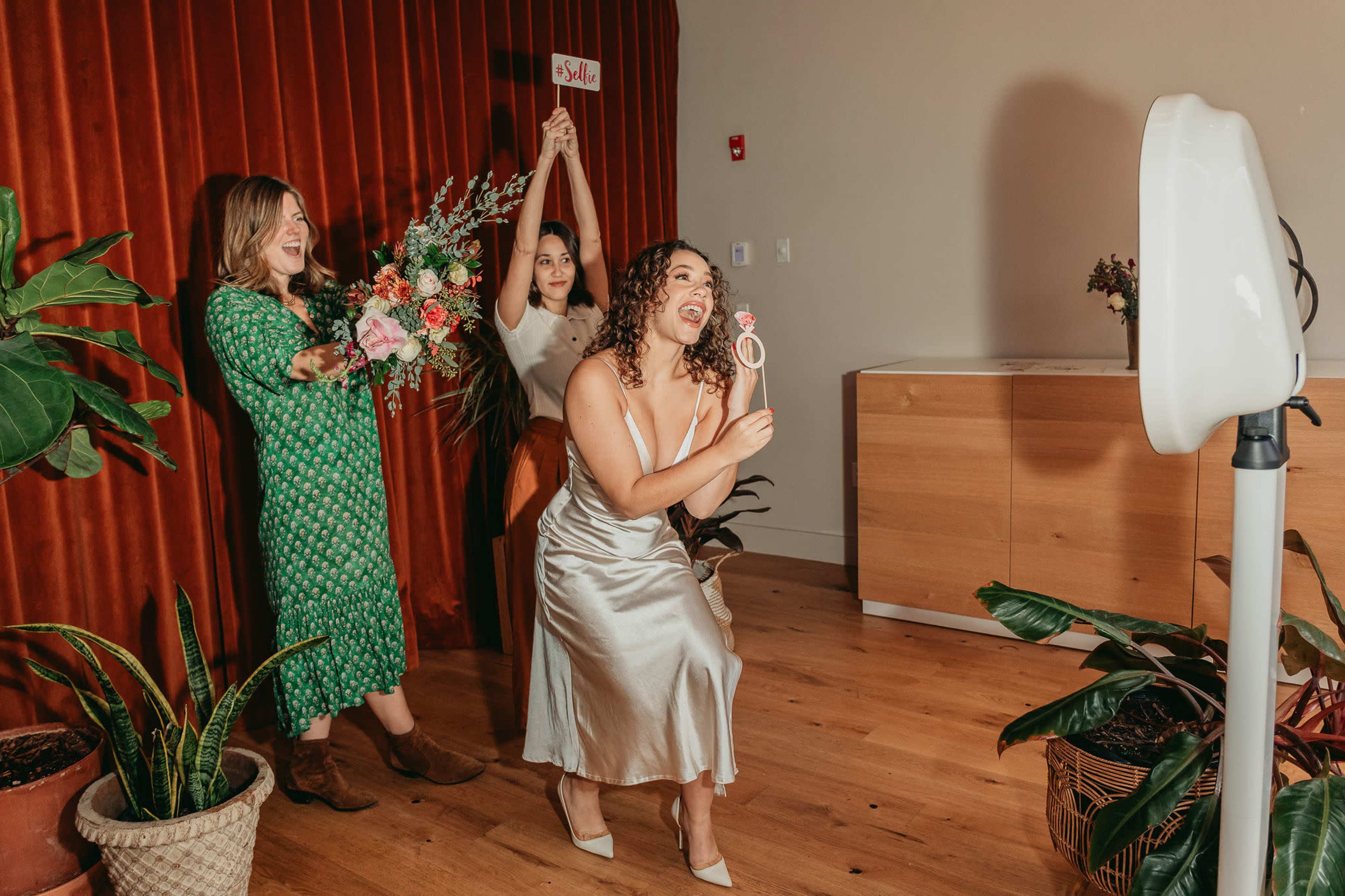 Three women pose in front of a photo booth, with one holding a bouquet and another holding a prop, while a third stands in a dress and smiles at the camera.