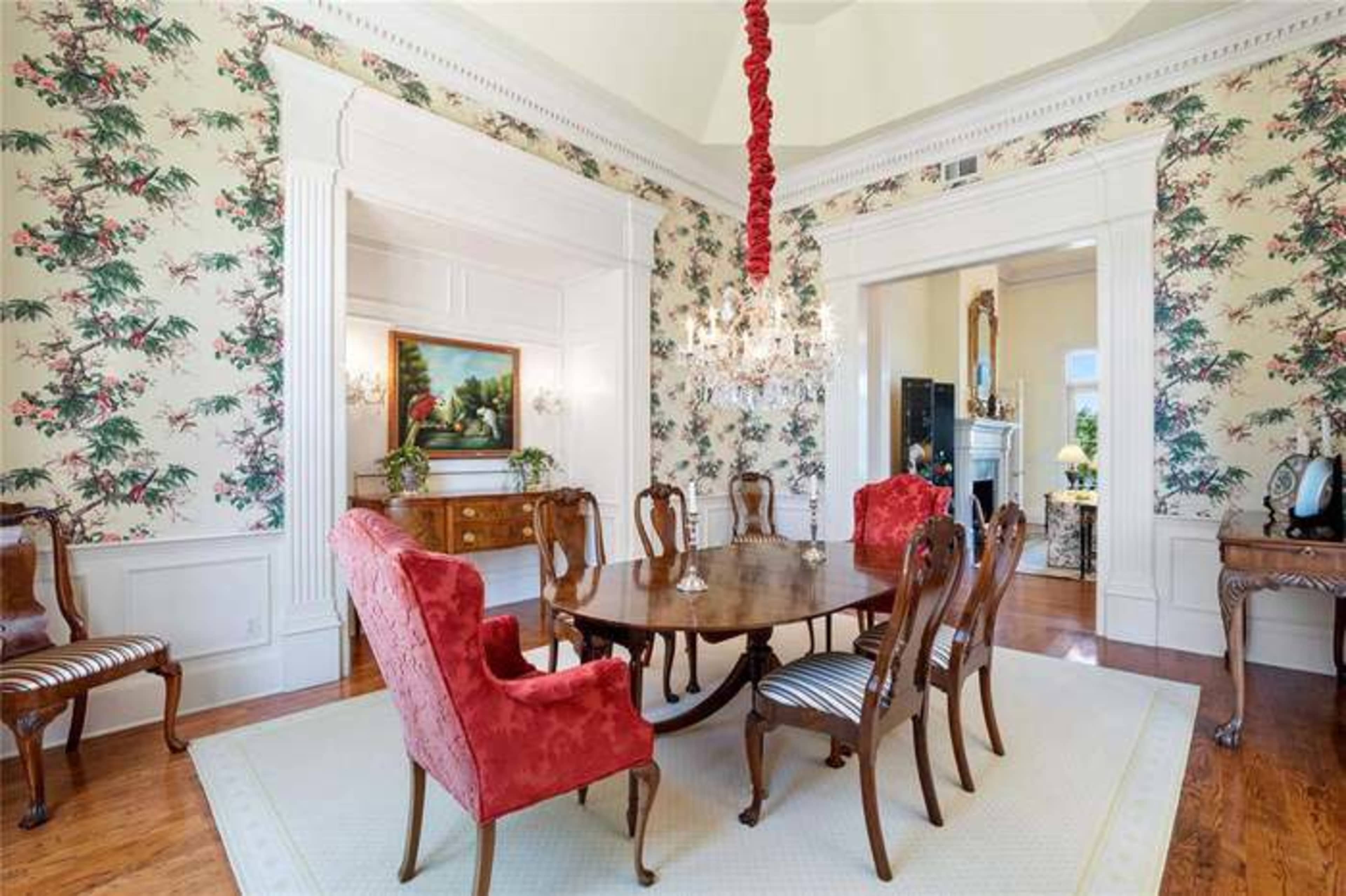 The dining room features a round wooden table surrounded by six upholstered chairs, with floral wallpaper and a chandelier overhead.