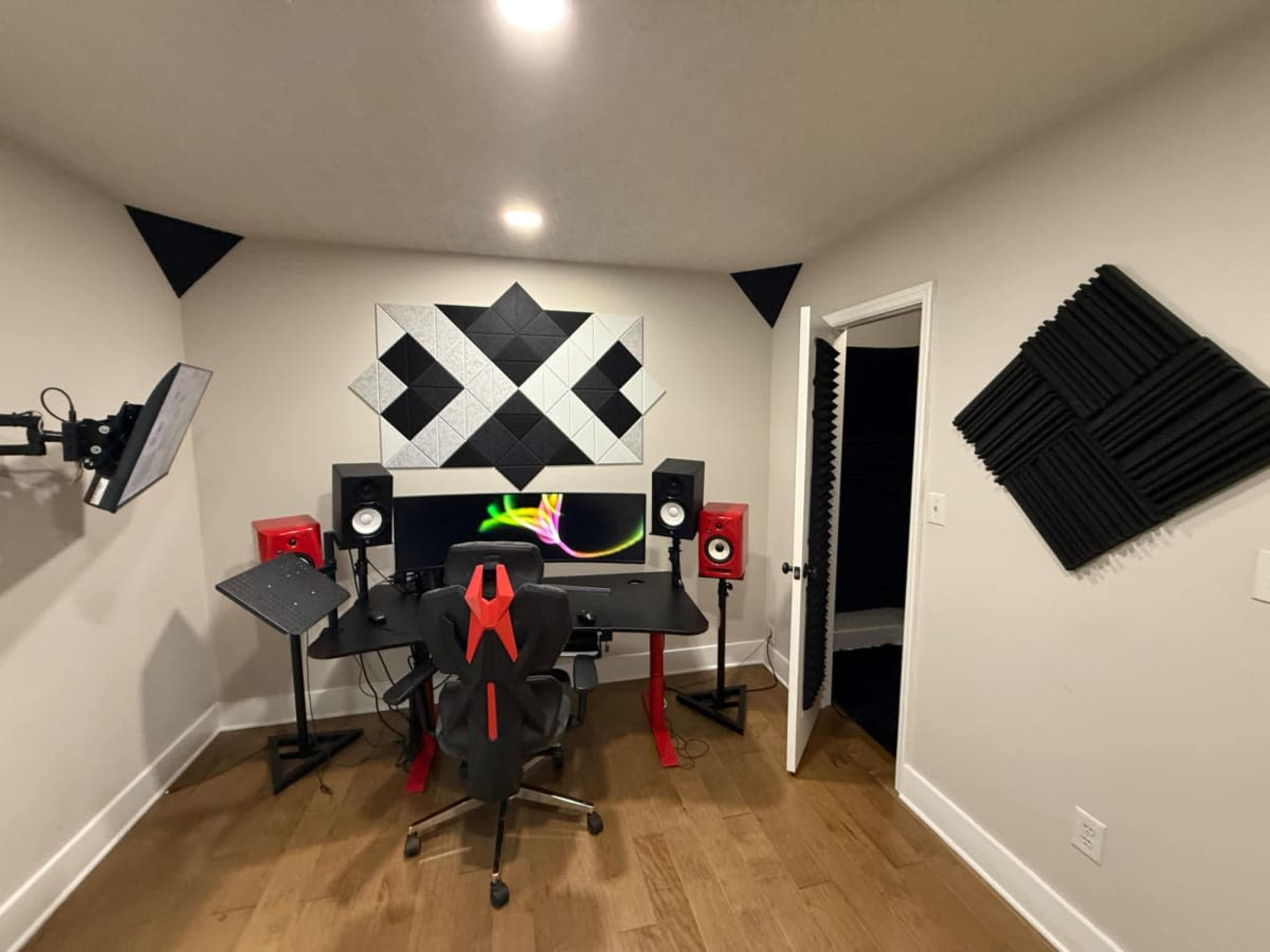 The image shows a modern recording studio with a black and red desk setup, featuring monitors and acoustic panels on the walls.