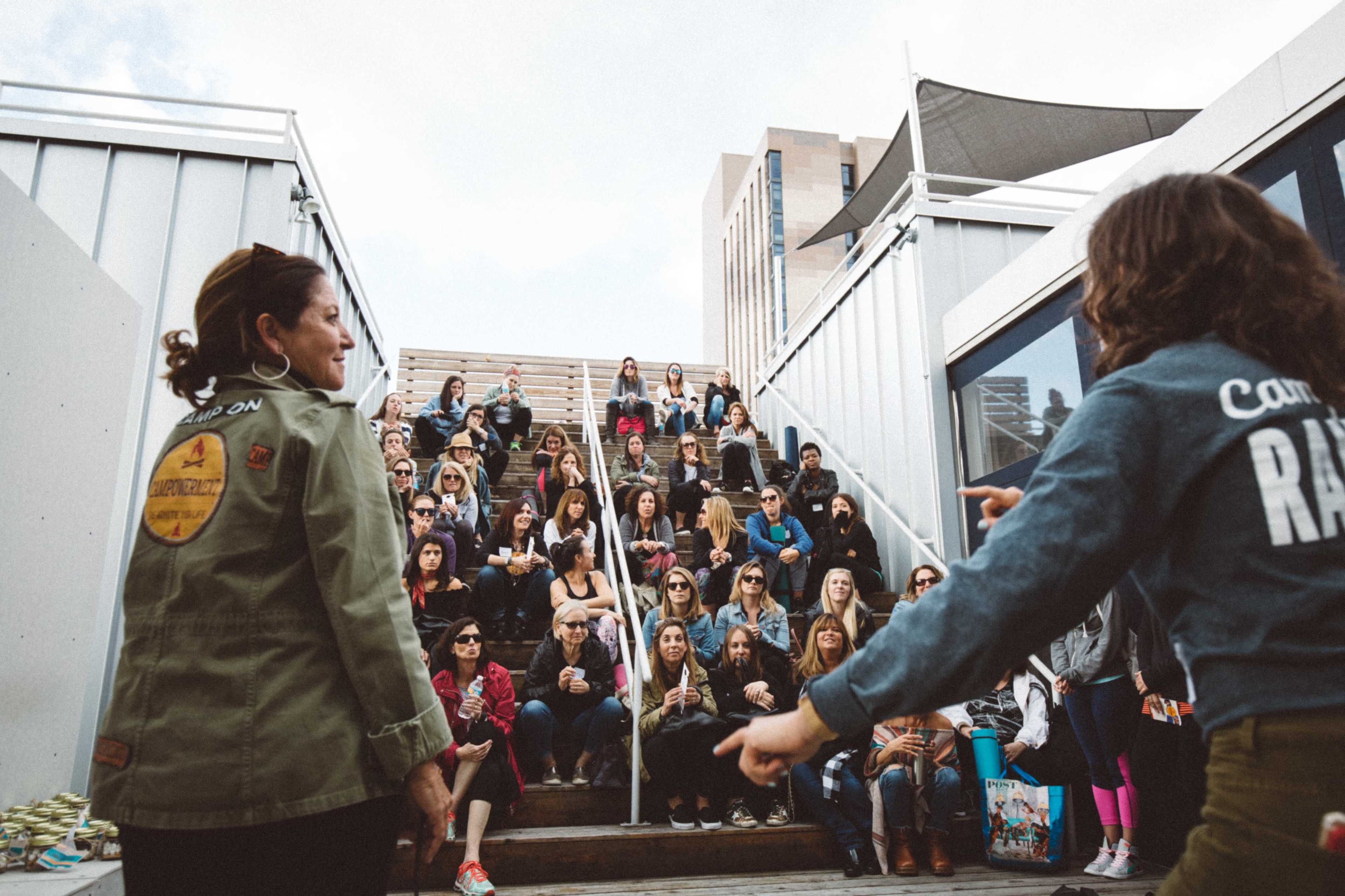 A crowd of people is seated on steps while two women stand at the front, engaging with the audience.