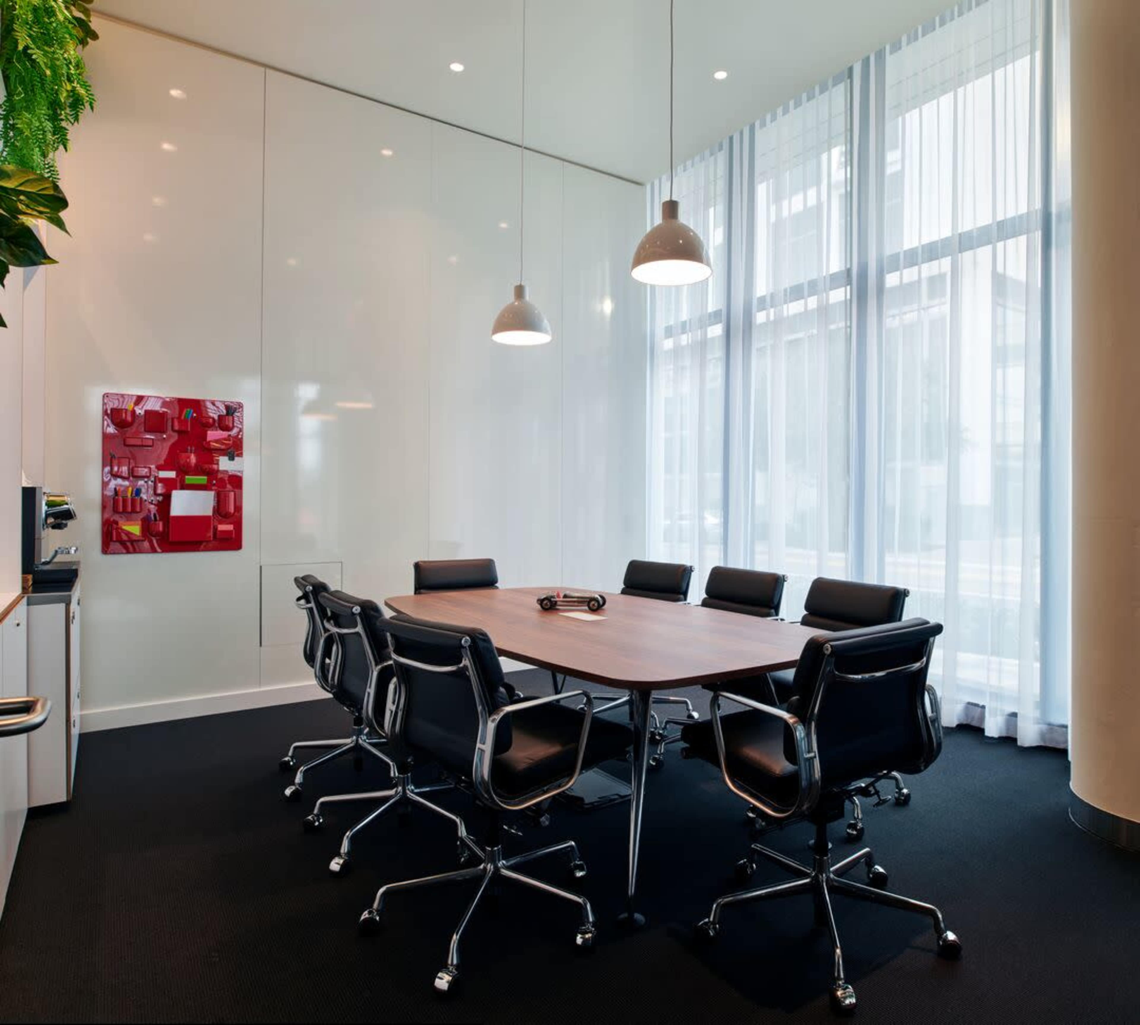 A conference room with a large wooden table surrounded by black leather chairs, illuminated by pendant lights, and featuring floor-to-ceiling curtains.