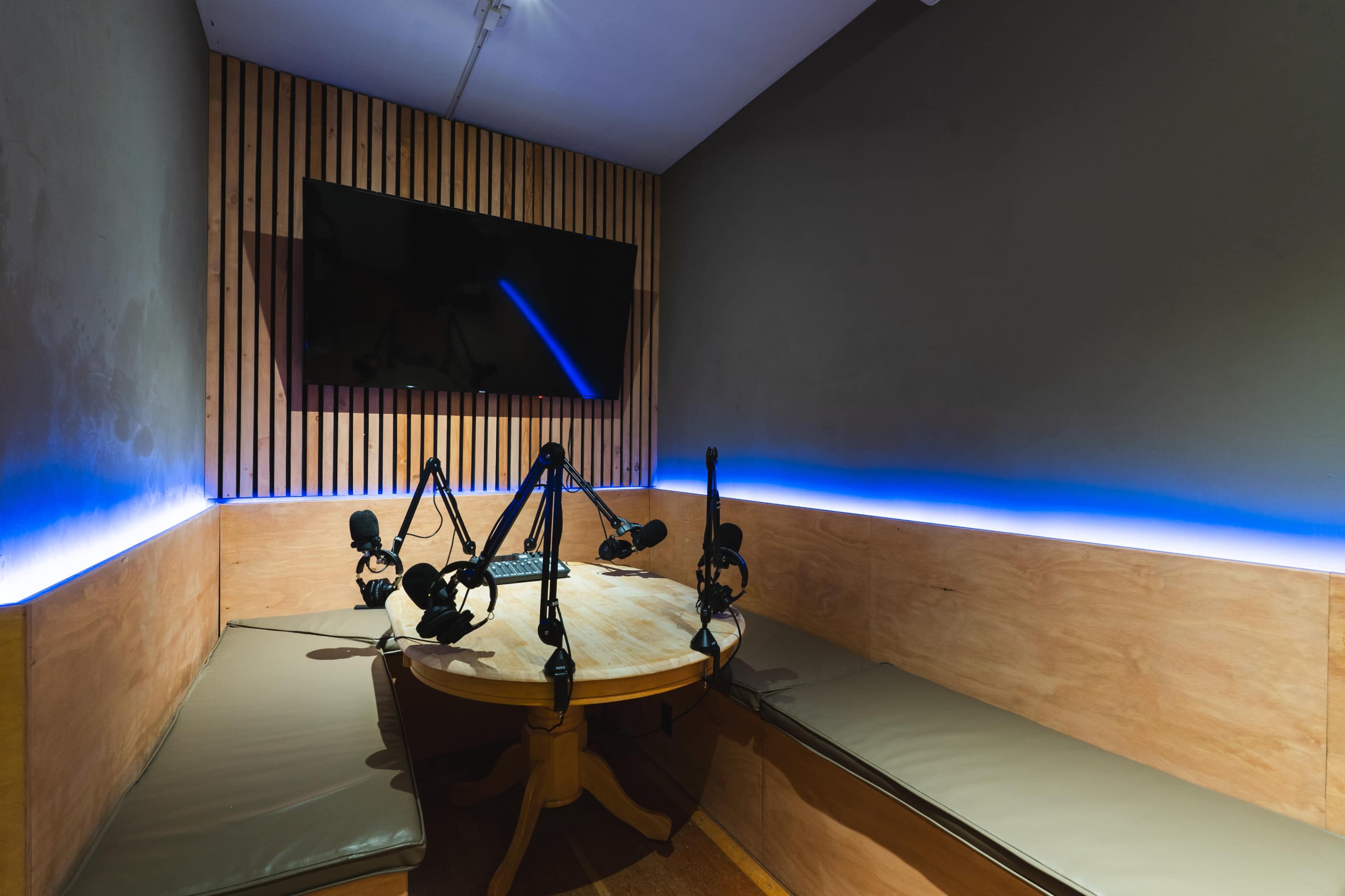 A small recording studio features a round table with seating, equipped with multiple microphones and a wall-mounted television.
