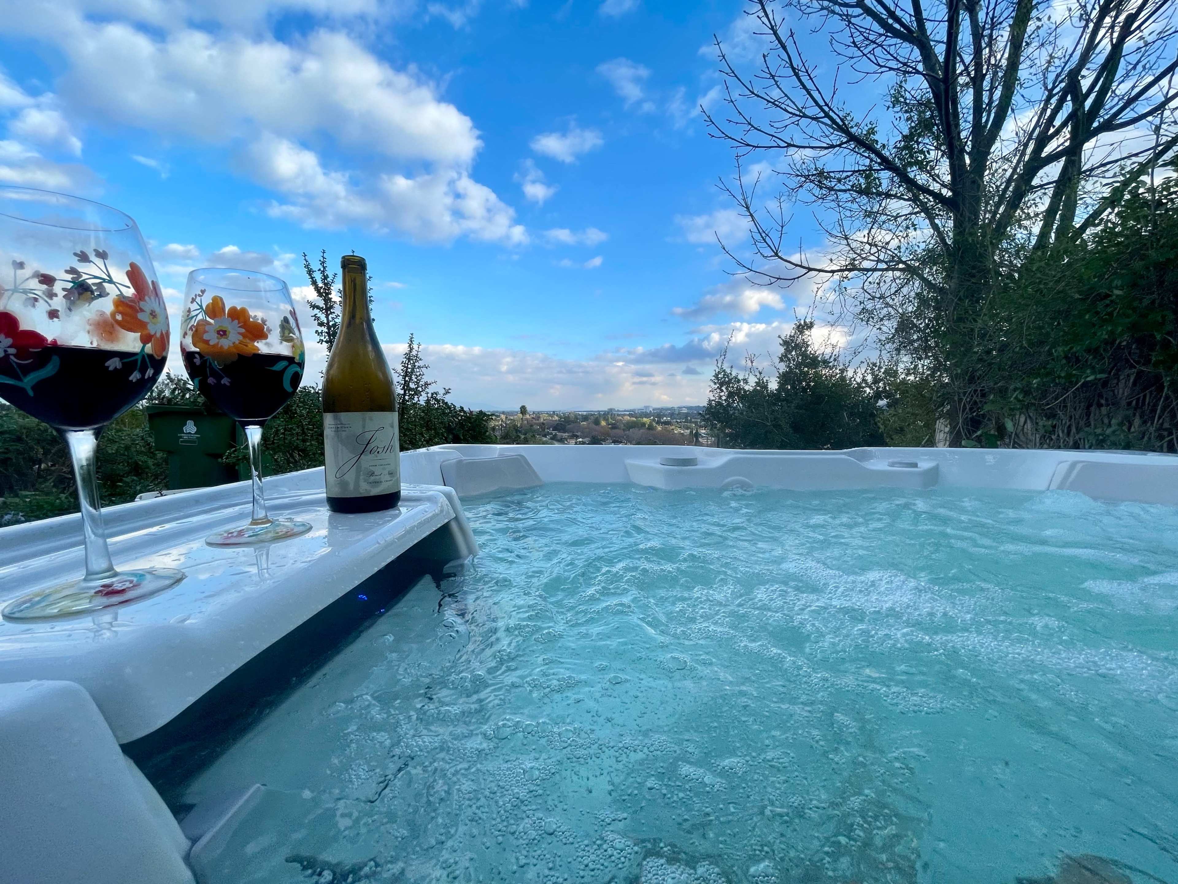 Two glasses of red wine and a bottle of white wine sit beside a hot tub with bubbling water, overlooking a landscape under a partly cloudy sky.