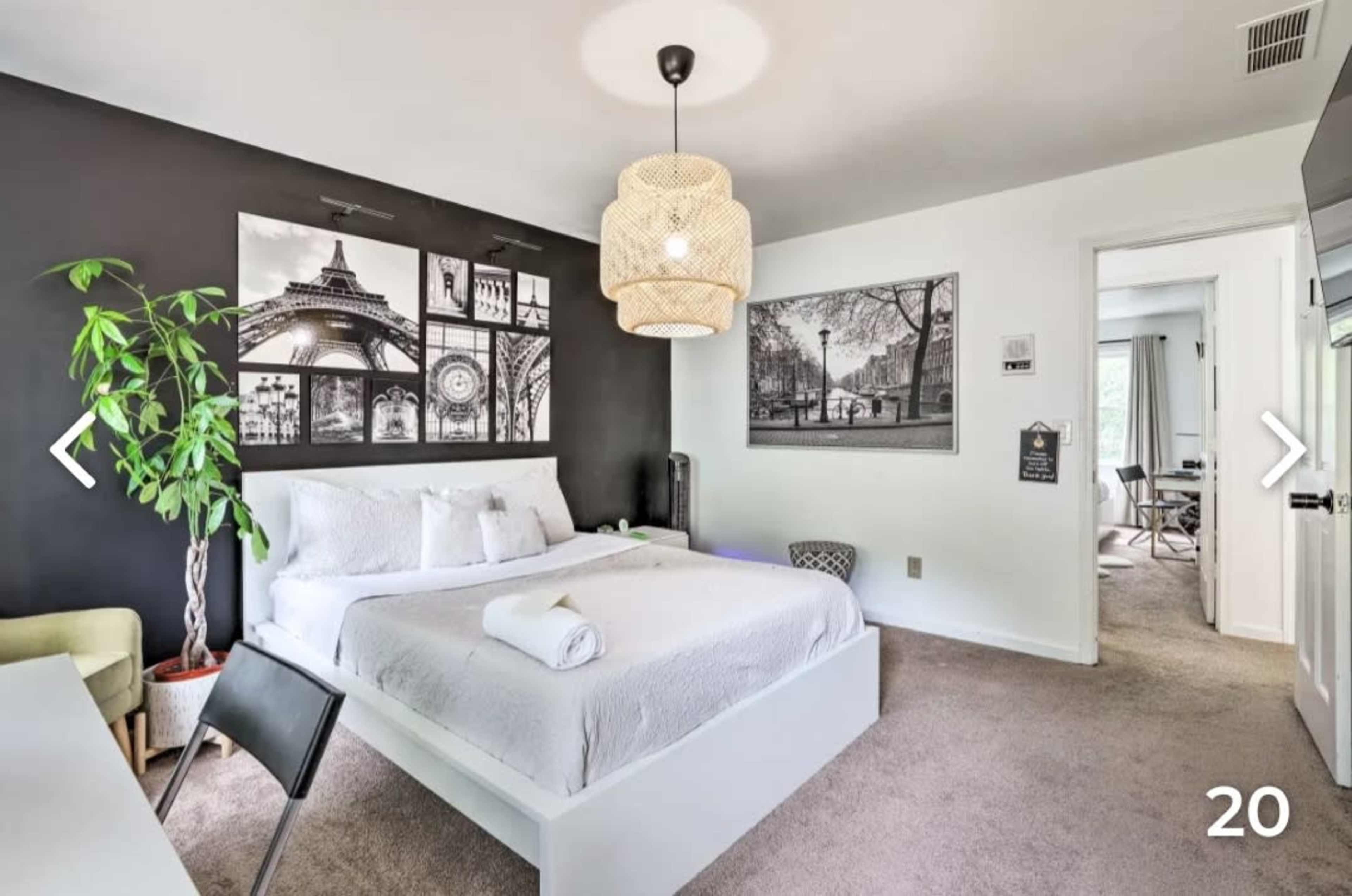 The image shows a modern bedroom featuring a large bed with white linens, a decorative wall with black and white artwork, and a potted plant beside a small desk and chair.