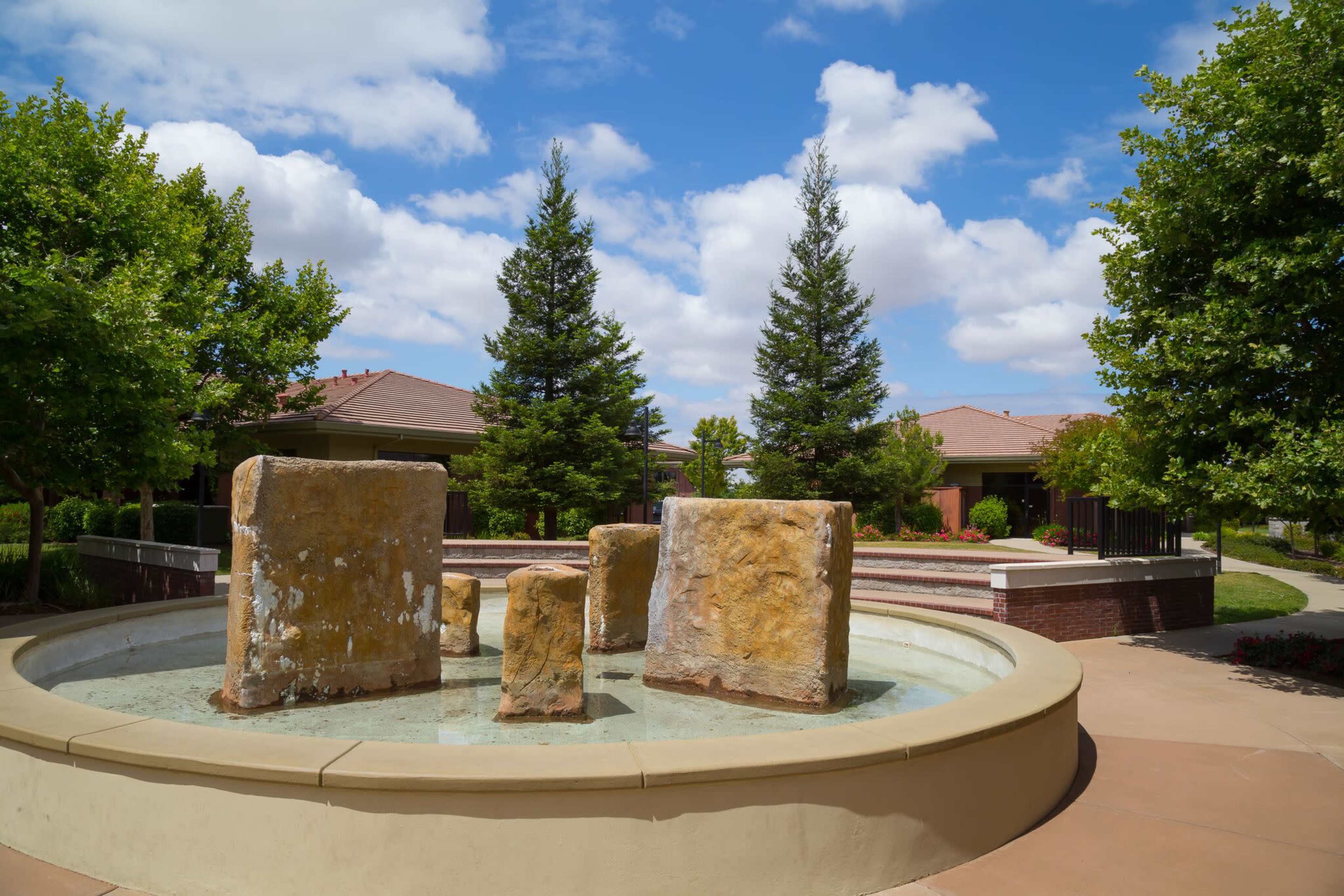 A fountain featuring large stone blocks sits in a landscaped area with green trees and residential buildings in the background.