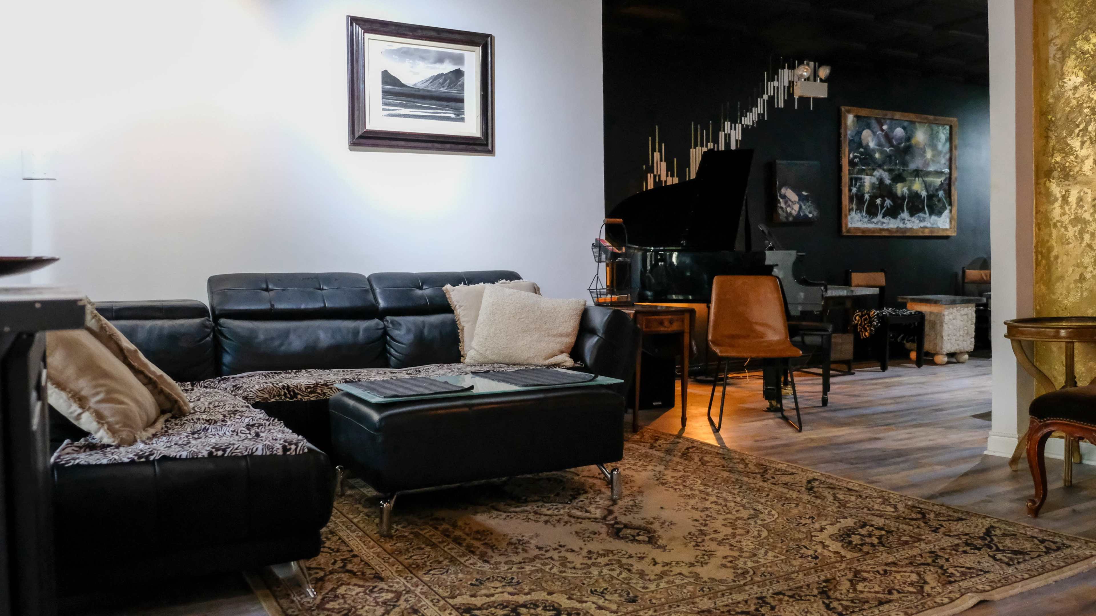 The image shows a modern living room with a black sectional sofa, an area rug, a grand piano in the background, and a mix of artwork and seating arrangements.